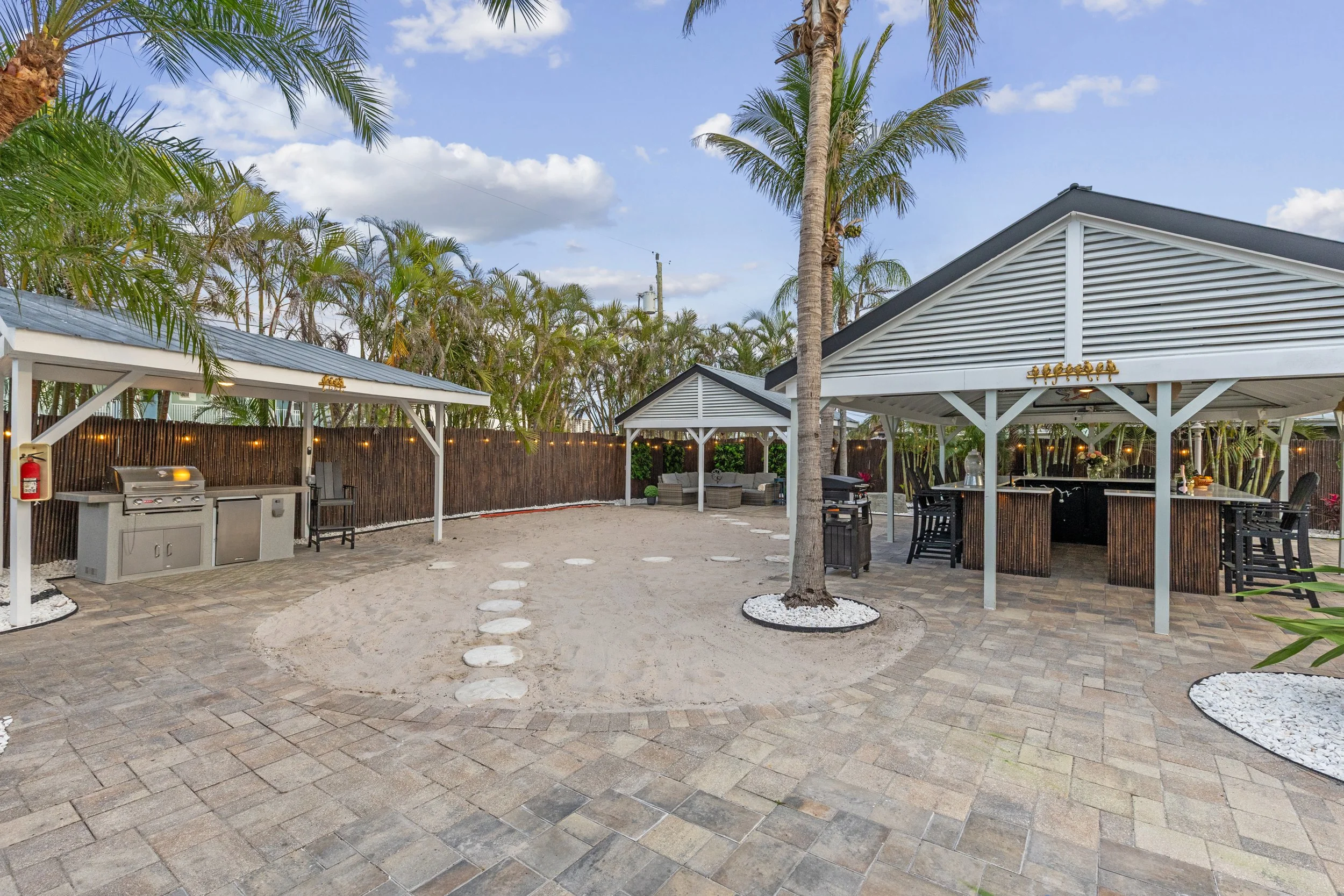 Outdoor backyard area with palm trees, sand, paved patio, covered bar area, sitting area with couches, and a grill, under a partly cloudy sky.
