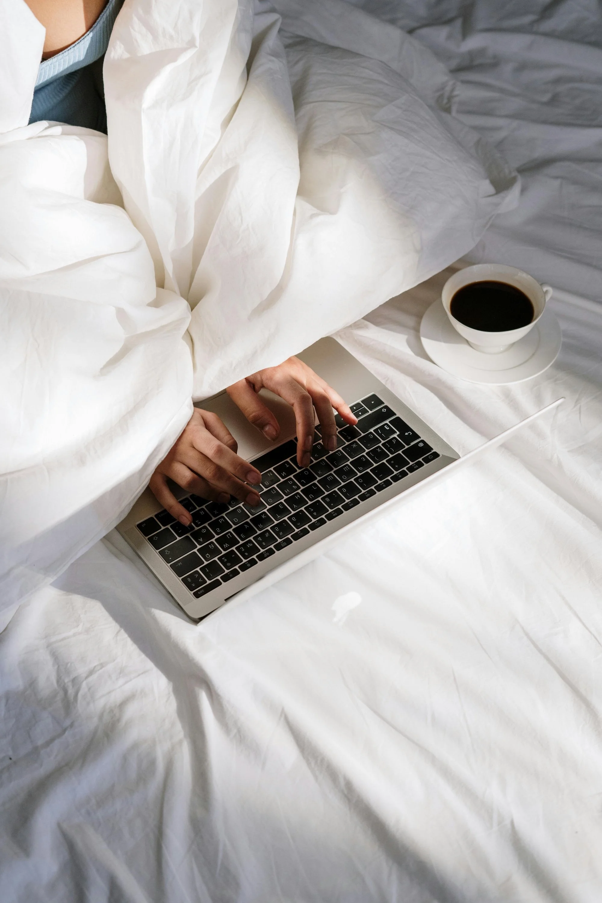 Person using a laptop on a bed with a cup of coffee nearby.
