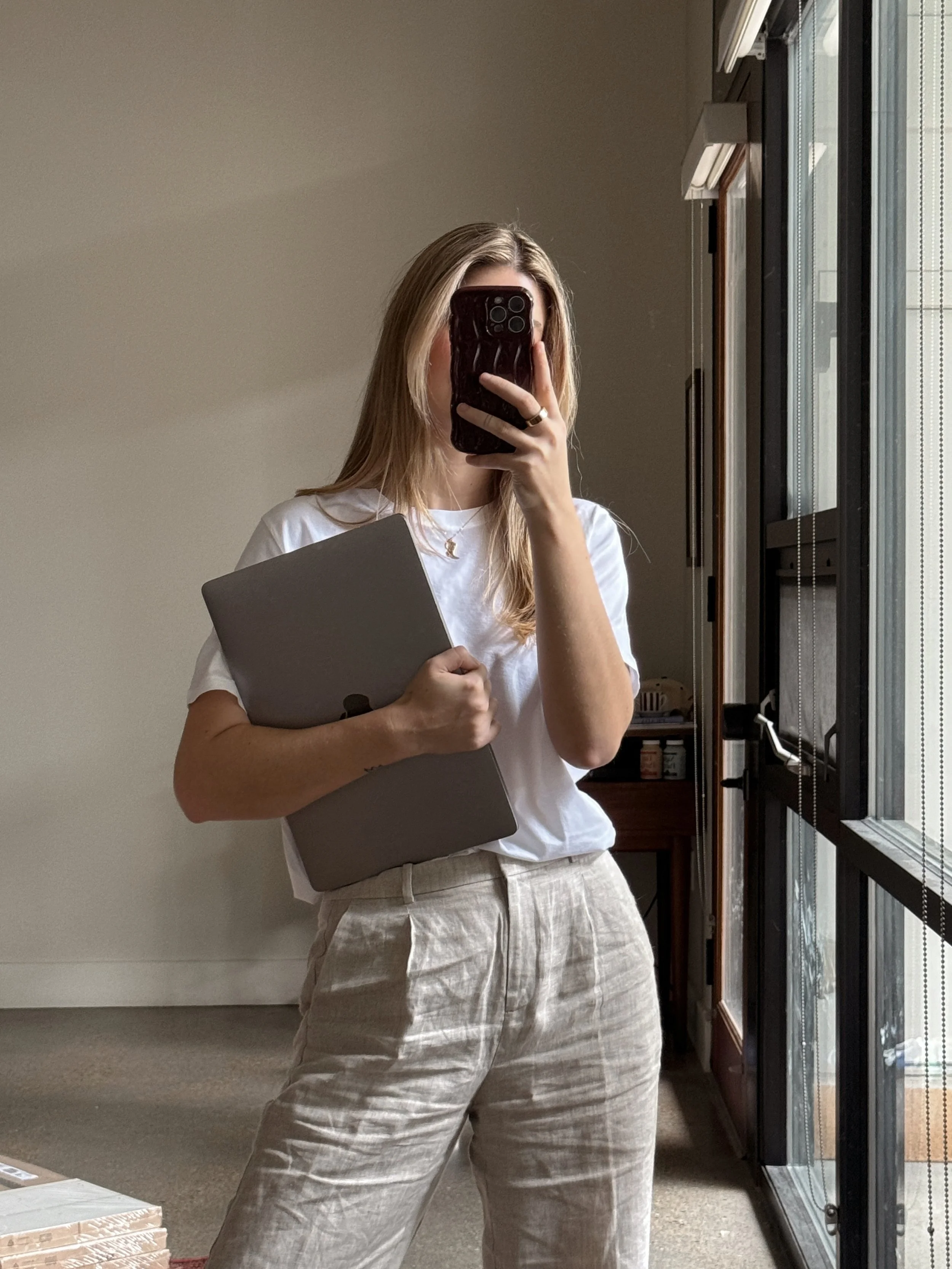 A woman with blonde hair taking a mirror selfie in a room, holding a gray folder or notebook and wearing a white T-shirt and beige pants.