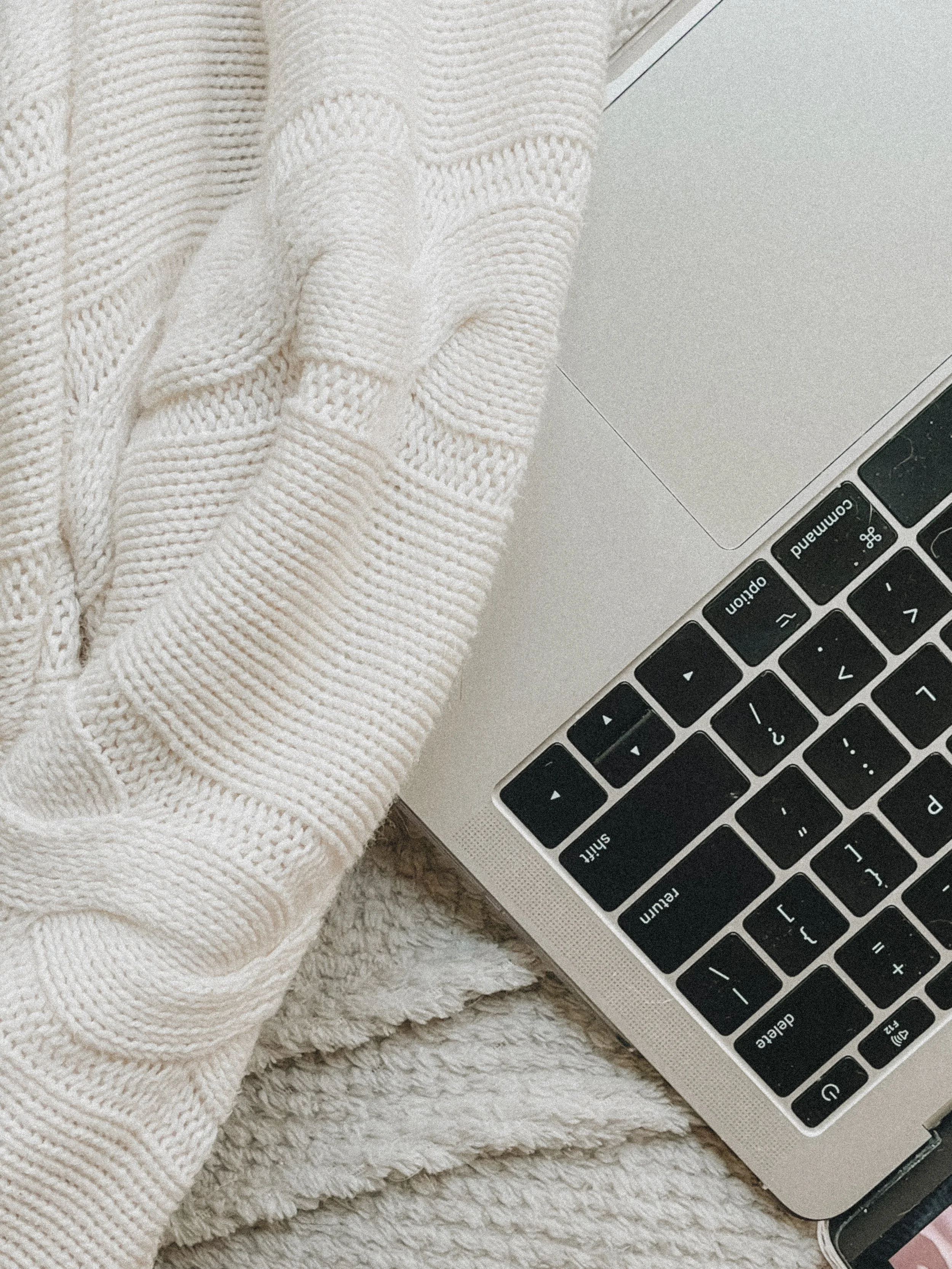 A person's hand resting on a laptop keyboard. The person is wearing a chunky white knit sweater. The laptop is silver with black keys, and the background includes a soft, textured blanket.
