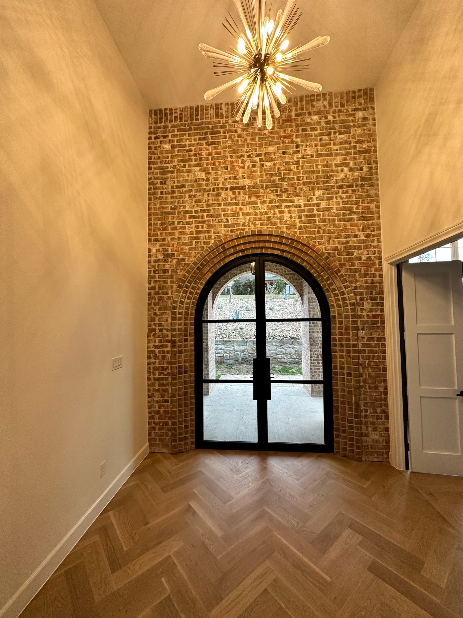 Interior view of a room with a brick wall, arched black metal door, wooden herringbone flooring, off-white walls, and a modern starburst chandelier hanging from the ceiling.