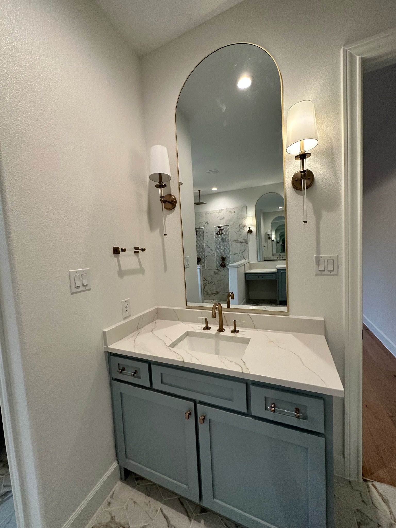 Bathroom vanity with a marble countertop, blue cabinetry, a large arched mirror, and brass hardware, with wall-mounted sconces on either side.