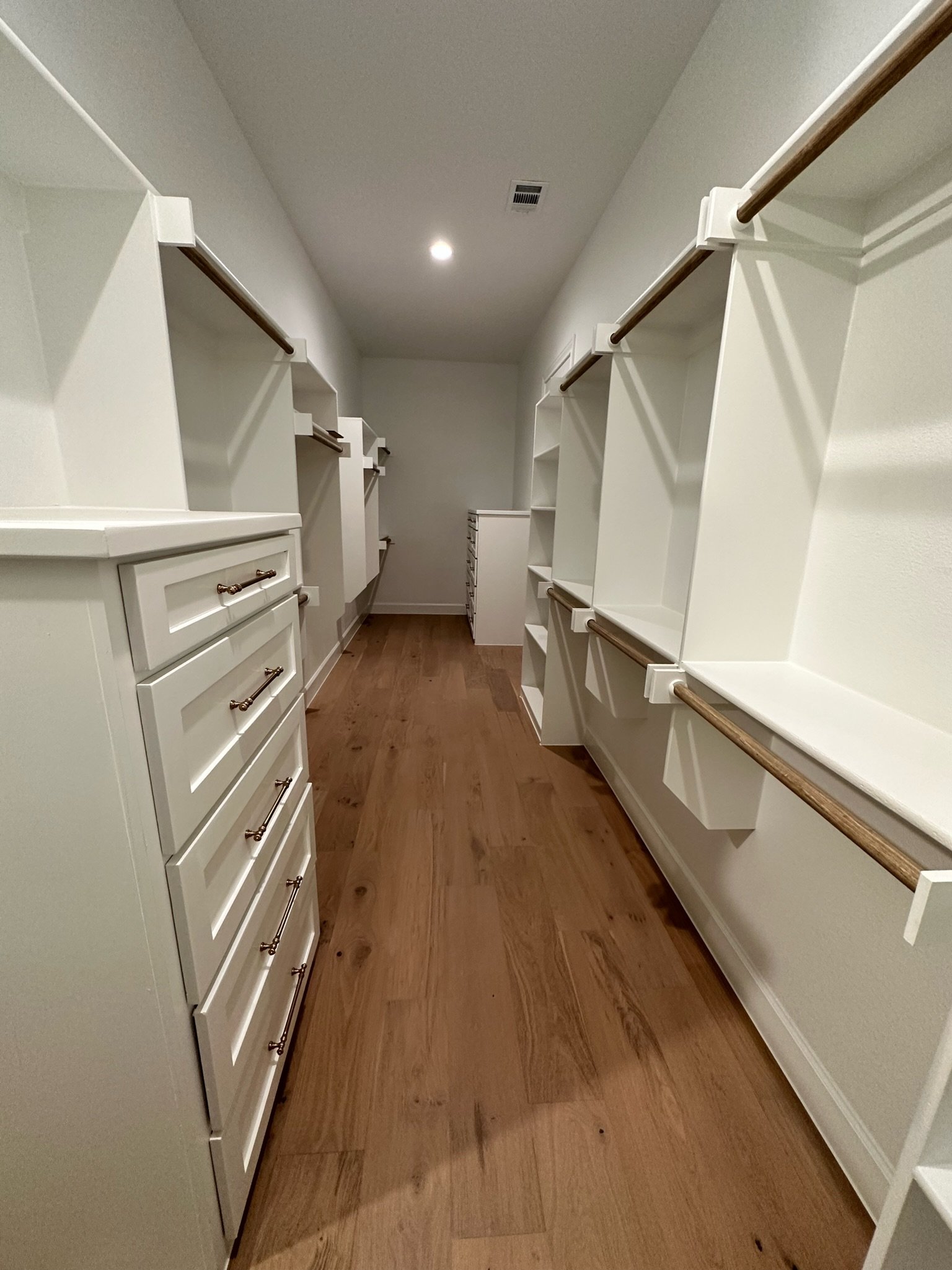Empty walk-in closet with white shelves and drawers, wooden rods for hanging clothes, and hardwood flooring.