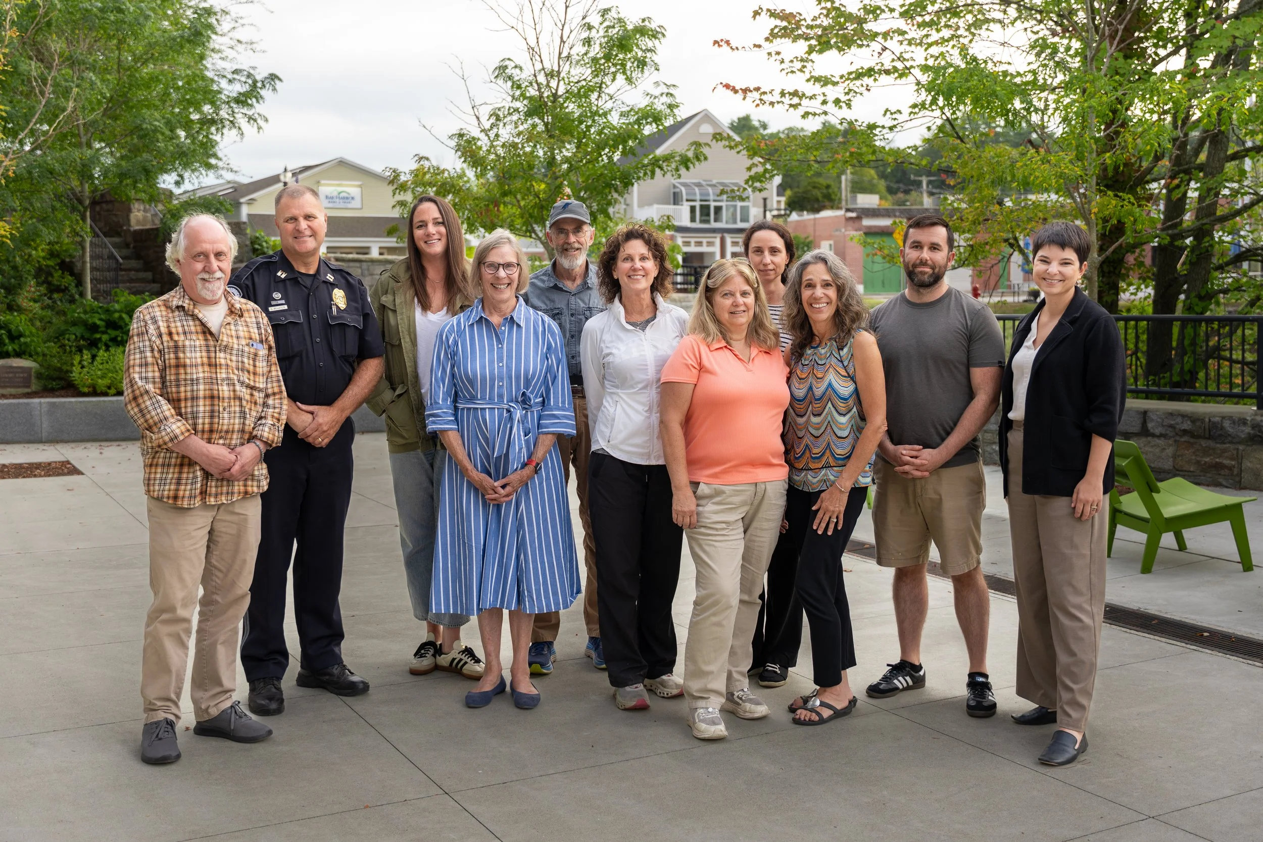 Group of twelve diverse people standing together outdoors in a park area, smiling at the camera, with trees and buildings in the background.