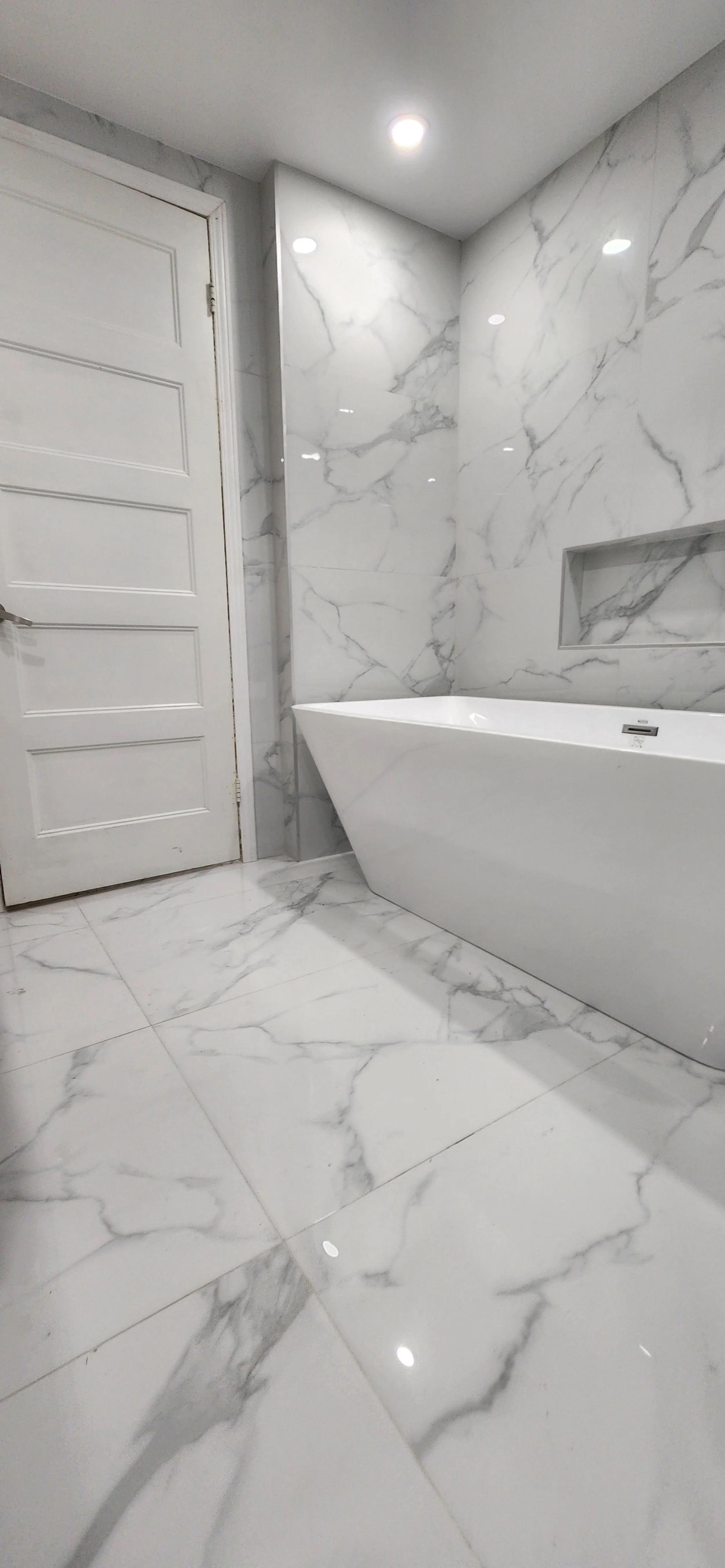 Modern bathroom with white marble walls and floor, a white bathtub, and a white door.