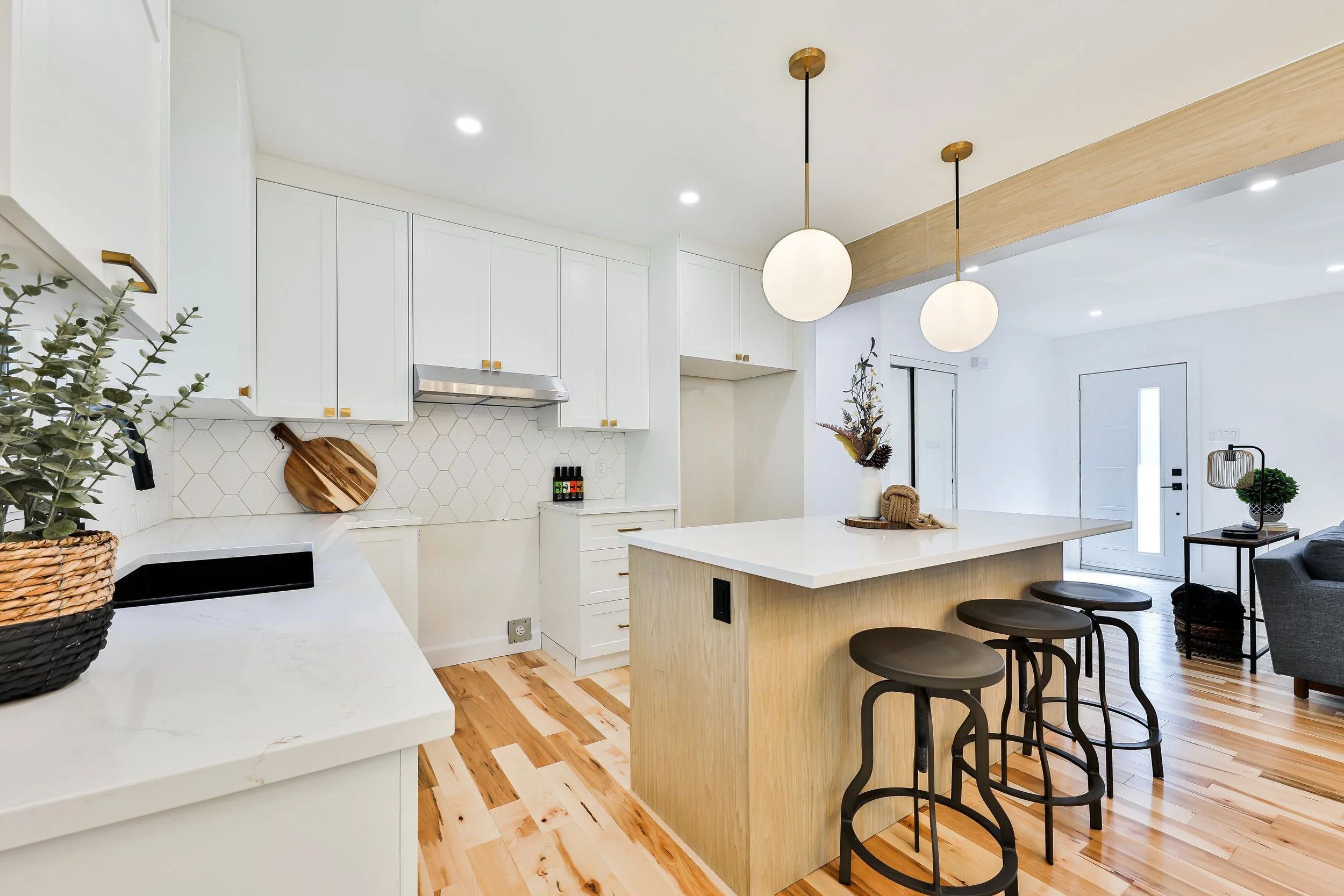 Modern open-concept kitchen with white cabinets, an island with a light wood finish, three black barstools, and pendant lights. The area features wooden flooring, a geometric backsplash, and minimal decor.
