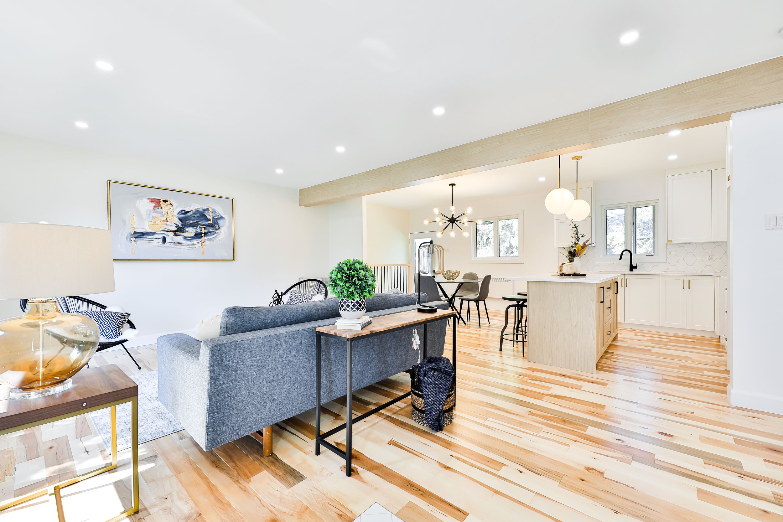 Open-concept living room and kitchen with hardwood floors, contemporary decor, a gray sofa, a black chair with a patterned pillow, a wooden side table with a lamp, a large abstract painting, a dining table with six chairs, modern lighting fixtures, w