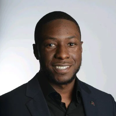 A professional headshot of a smiling man with dark skin, wearing a dark suit and black shirt, against a plain light background.