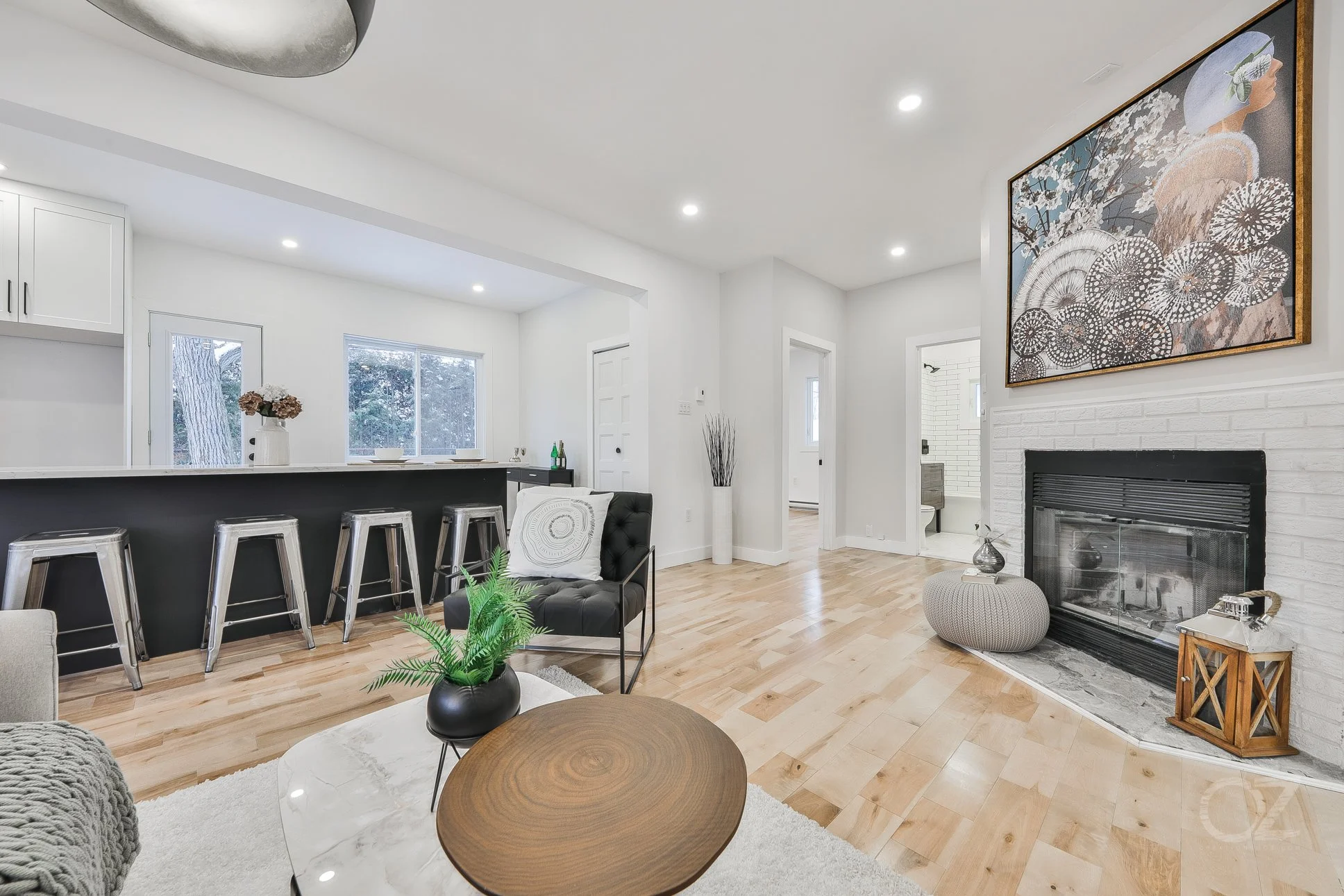 Modern living room with a white brick fireplace, abstract artwork above, light wooden floor, black accent sofa, marble-top coffee table with a black vase and green plant, bar area with four metallic barstools, white walls and ceiling, windows, and de
