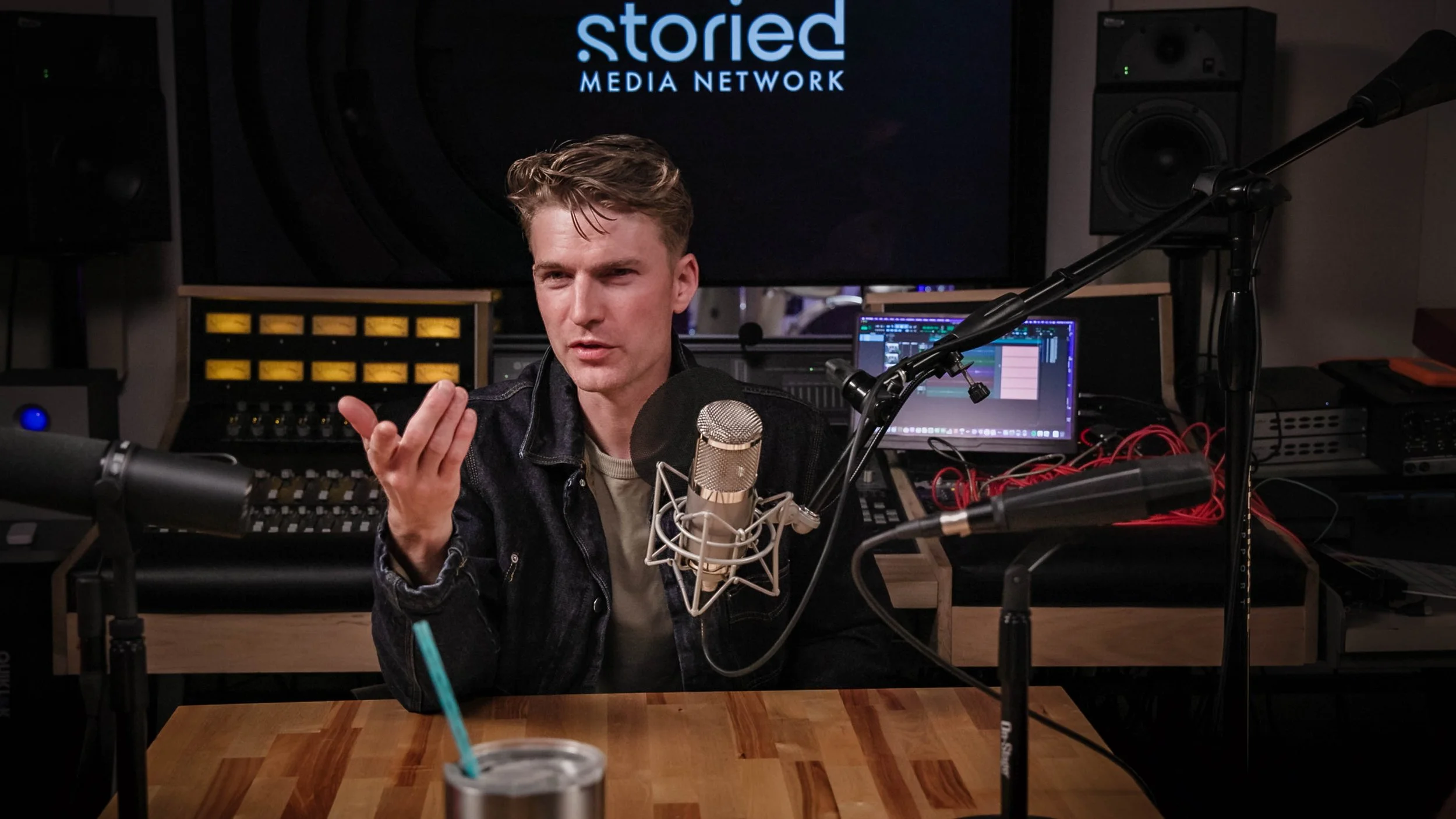 Chris Zobac sitting in the control room of Three21 studios with microphones, audio equipment, and screens, under a sign reading 'storied media network.'