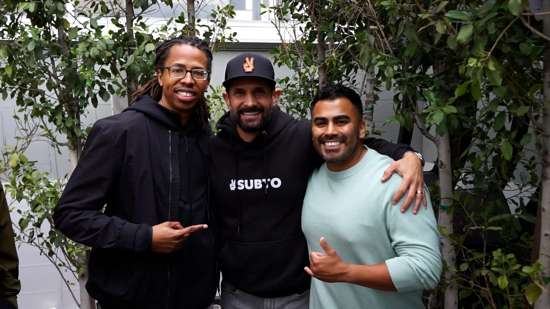 Three men smiling, standing outdoors in front of green foliage. The man on the left is wearing glasses and a black jacket, the man in the middle is wearing a black hoodie with a logo, and the man on the right is wearing a light green sweater. They are posing closely with arms around each other.