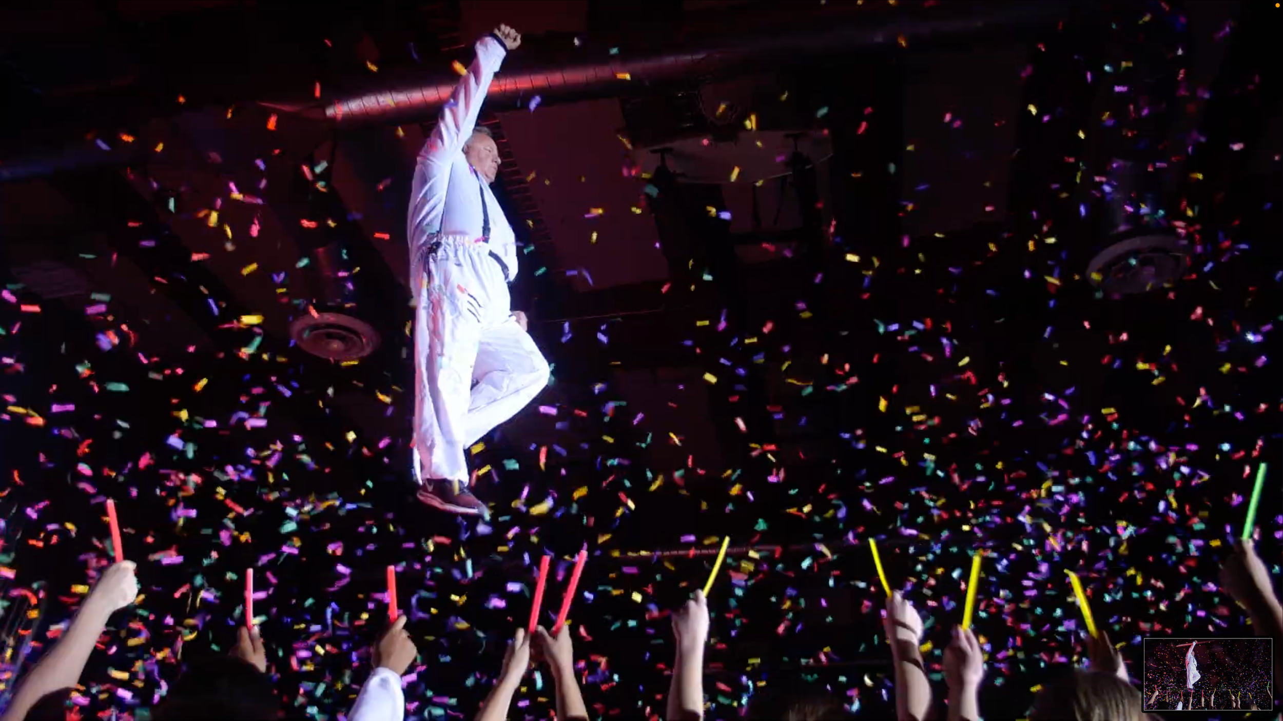 Performer in white outfit levitating on stage amid colorful confetti during a celebration with audience holding glow sticks.