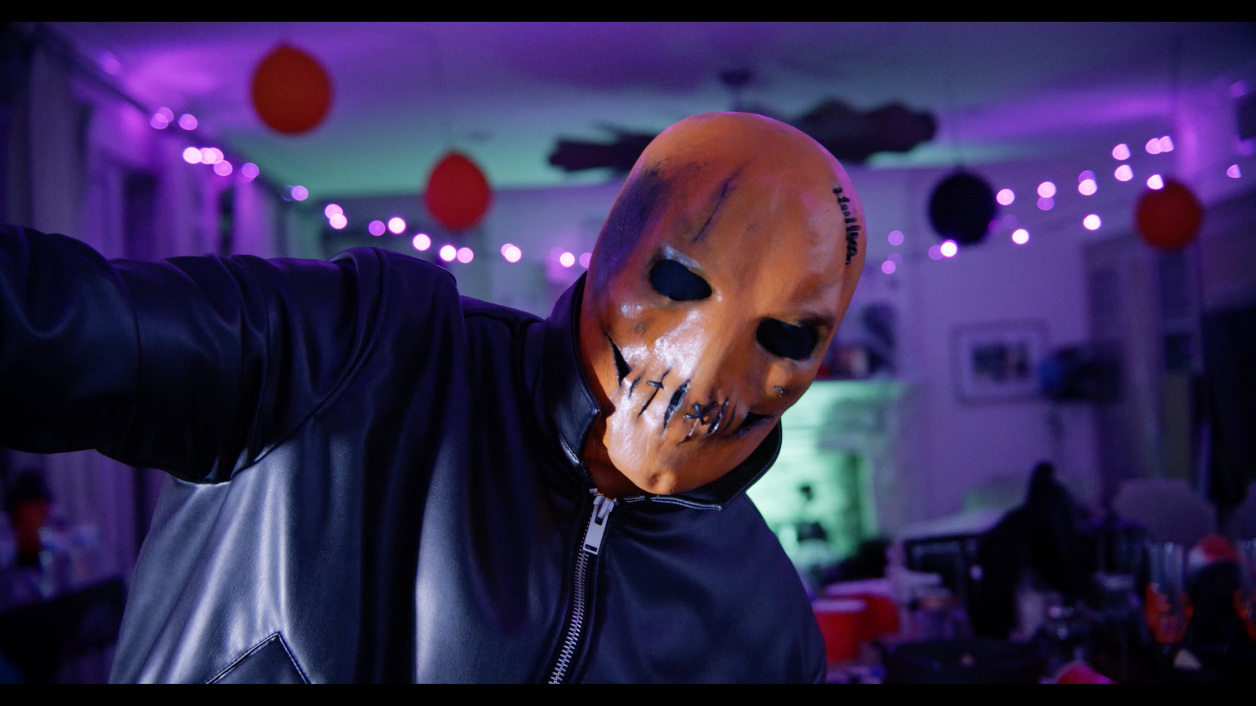 Person wearing a horror mask with black eye holes and stitched mouth, taking a selfie in a room decorated with purple string lights and red paper lanterns.