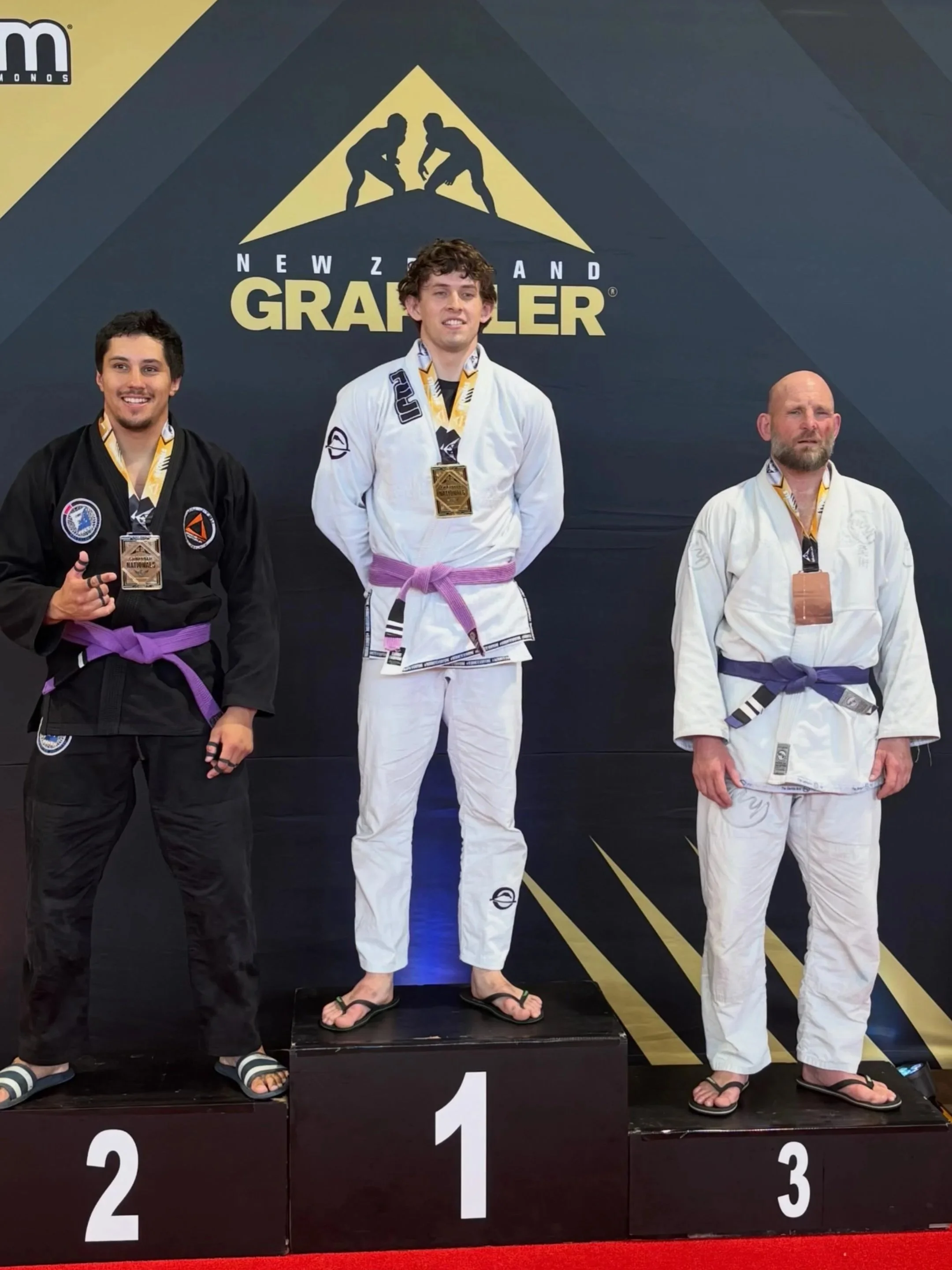 Three male martial artists in judo uniforms on a winners' podium with medals, standing in front of a black backdrop with a yellow logo featuring two judo fighters and the words 'New Zealand Grappler.'