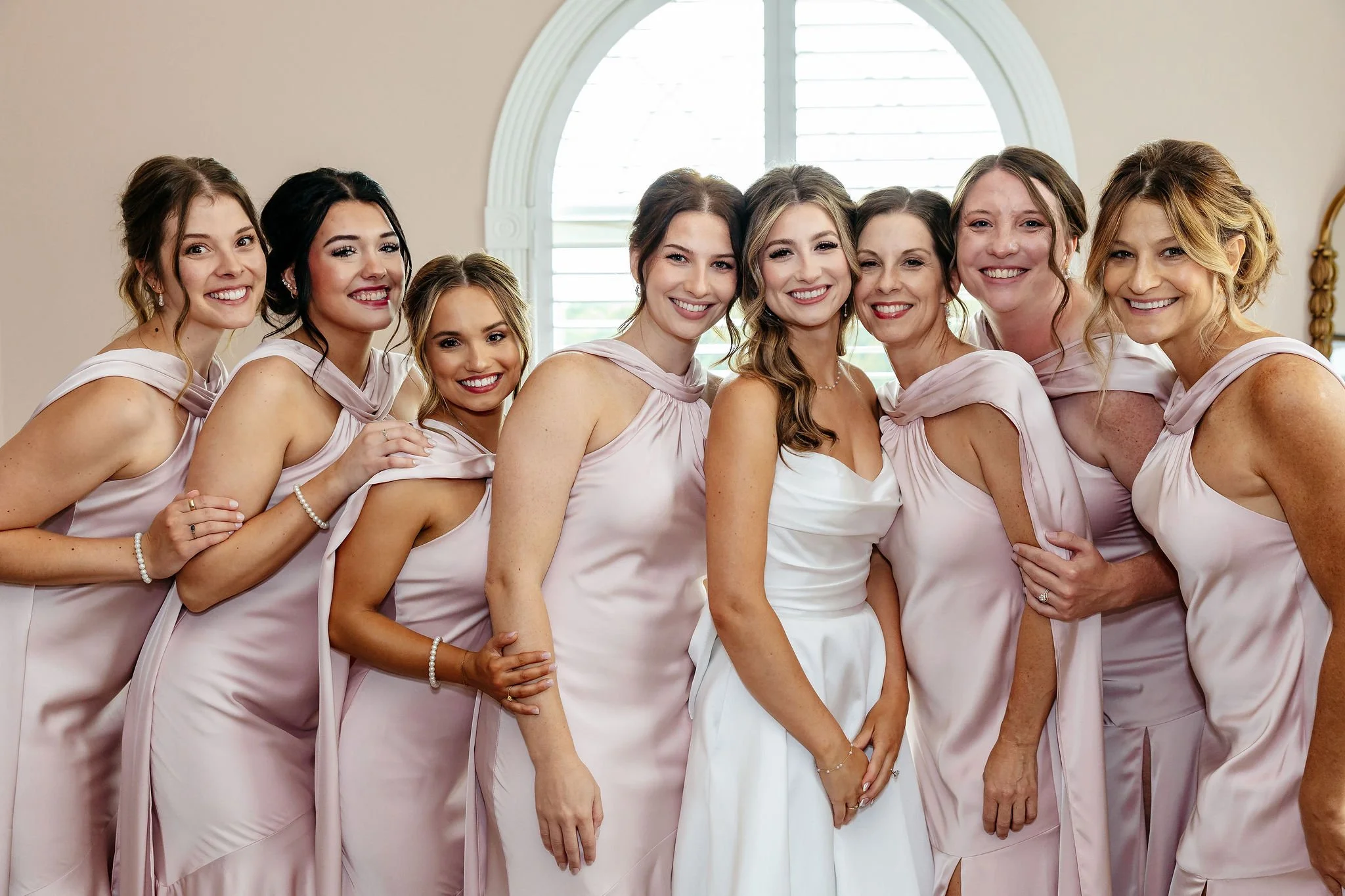 A group of eight women, including a bride in a white dress and seven bridesmaids in light pink dresses, smiling and posing together indoors with a window in the background.