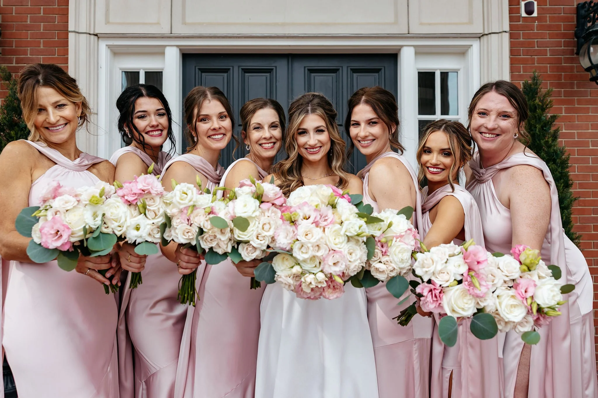 A bride and seven bridesmaids standing in front of a house, holding large bouquets of pink and white flowers, smiling at the camera.