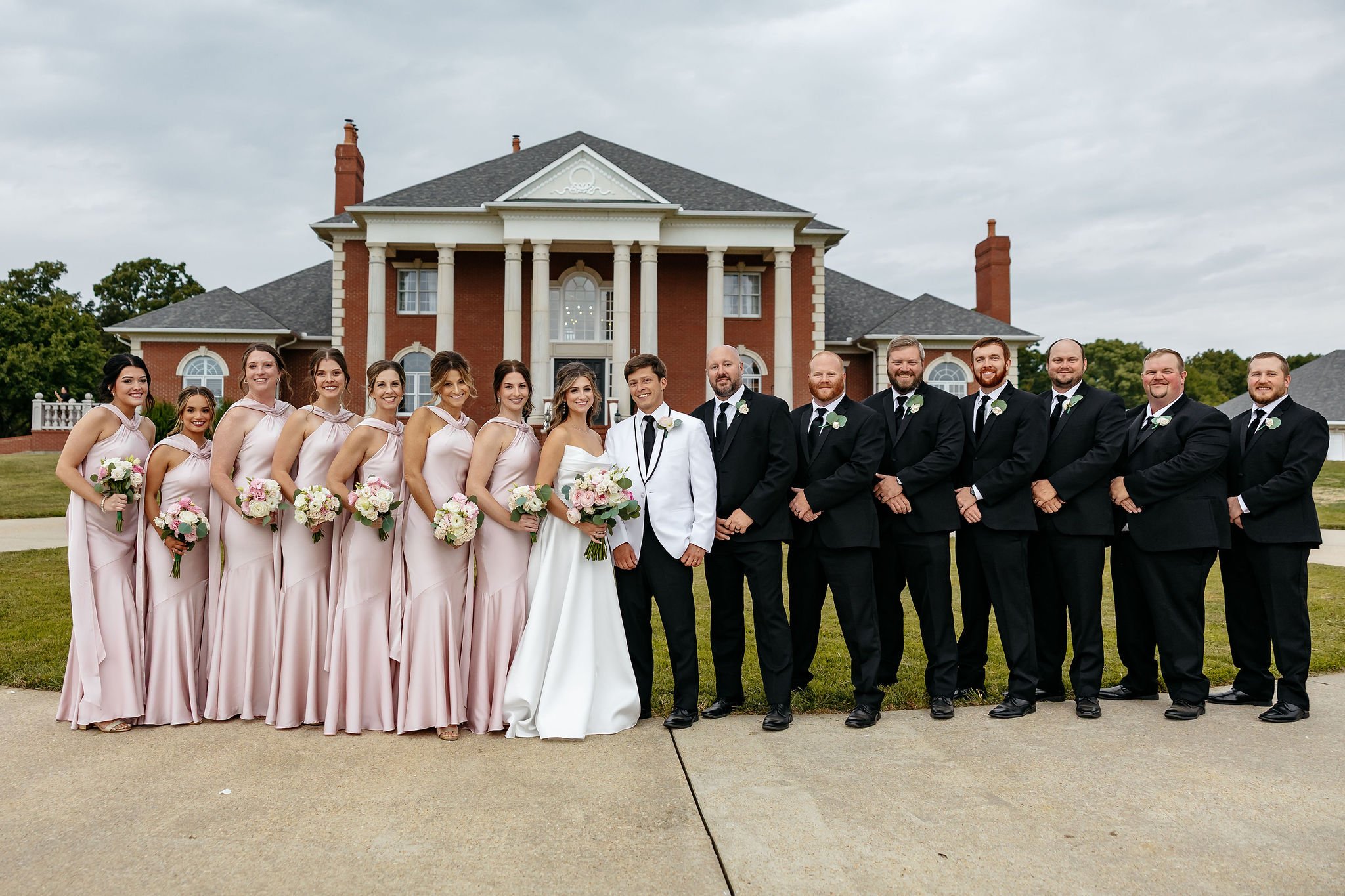 Wedding party standing in front of a large brick house with a portico. Bridesmaids wear pink dresses and hold bouquets, groomsmen wear black suits with white shirts and black ties, and the bride and groom stand in the center, smiling.