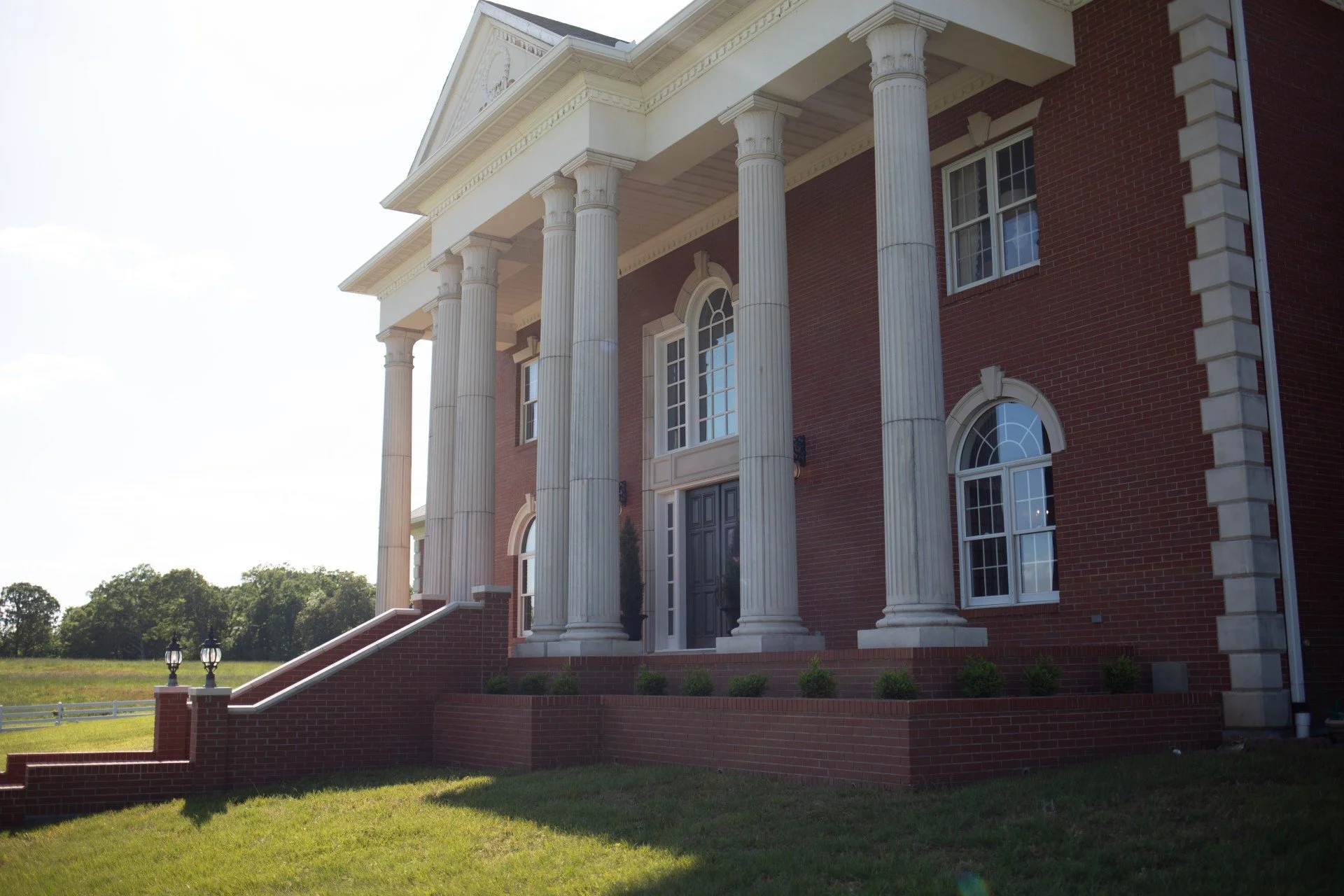 Front view of a large, red brick mansion with white classical columns supporting a front porch, and multiple windows with arched and rectangular shapes, surrounded by a grassy yard and trees in the background.