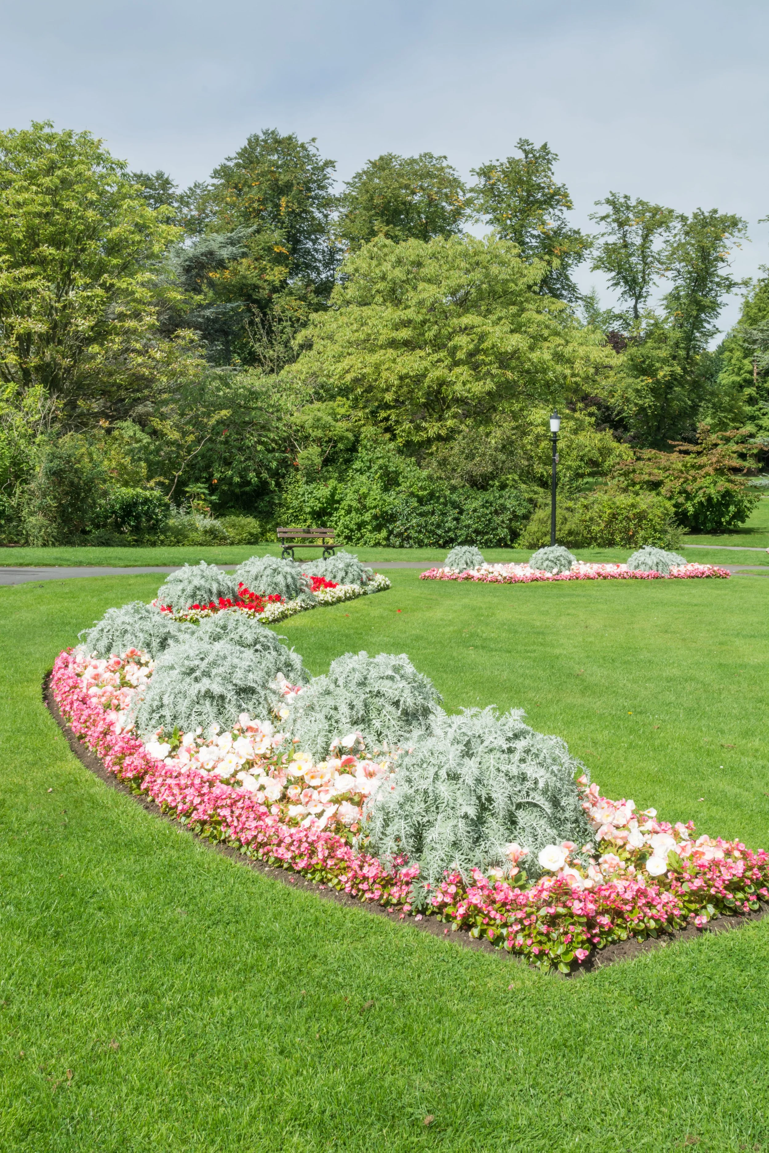A well-maintained park with a green lawn, colorful flower beds, a bench, a lamppost, and lush trees in the background under a clear sky.