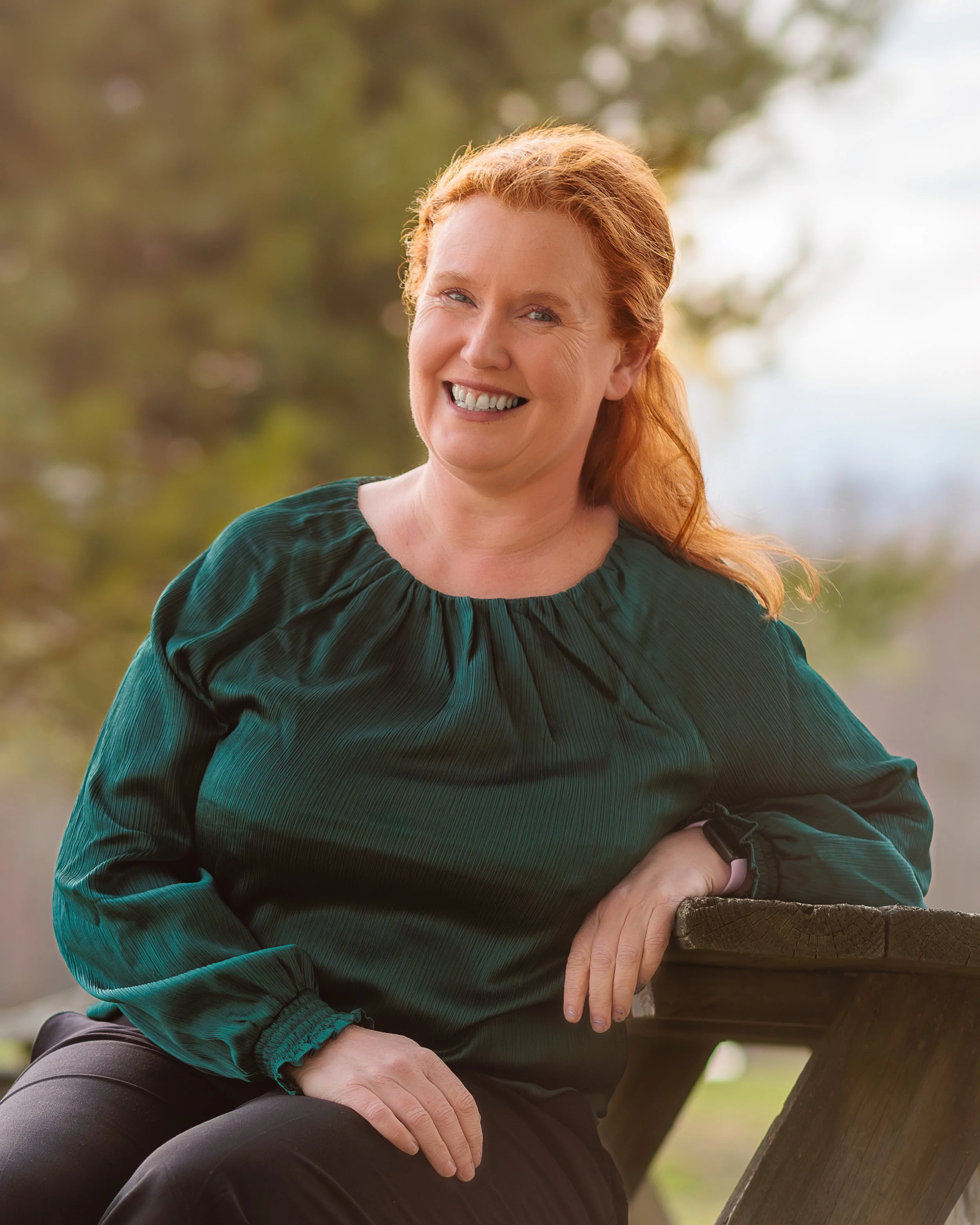 A woman with long red hair smiles warmly while seated outdoors, wearing a teal green blouse. Trees and soft natural light are visible in the background.