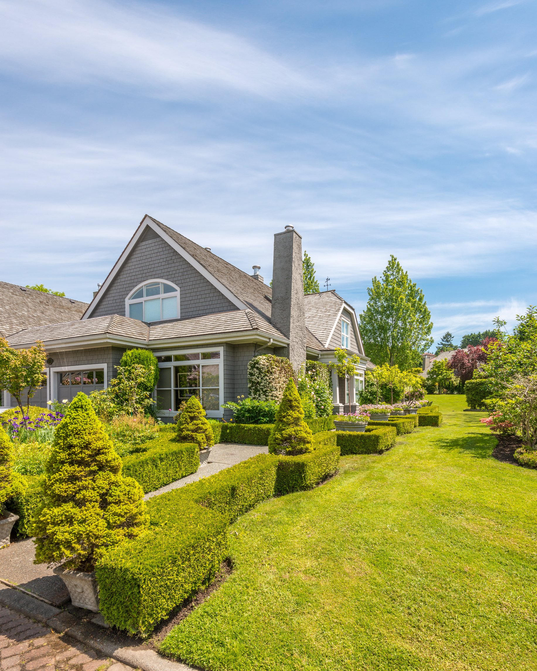Well-maintained front yard garden of a house with trimmed hedges, various flowering plants, and a lush green lawn, under a partly cloudy sky.
