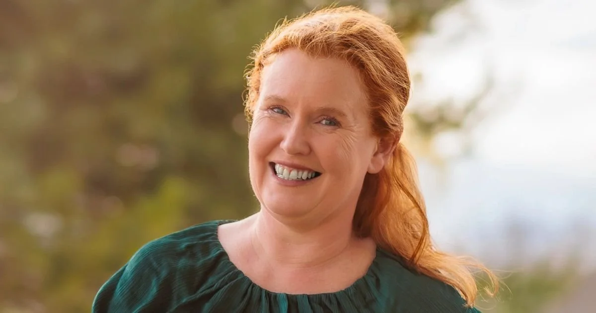 A woman with red hair smiling outdoors with blurred trees in the background.