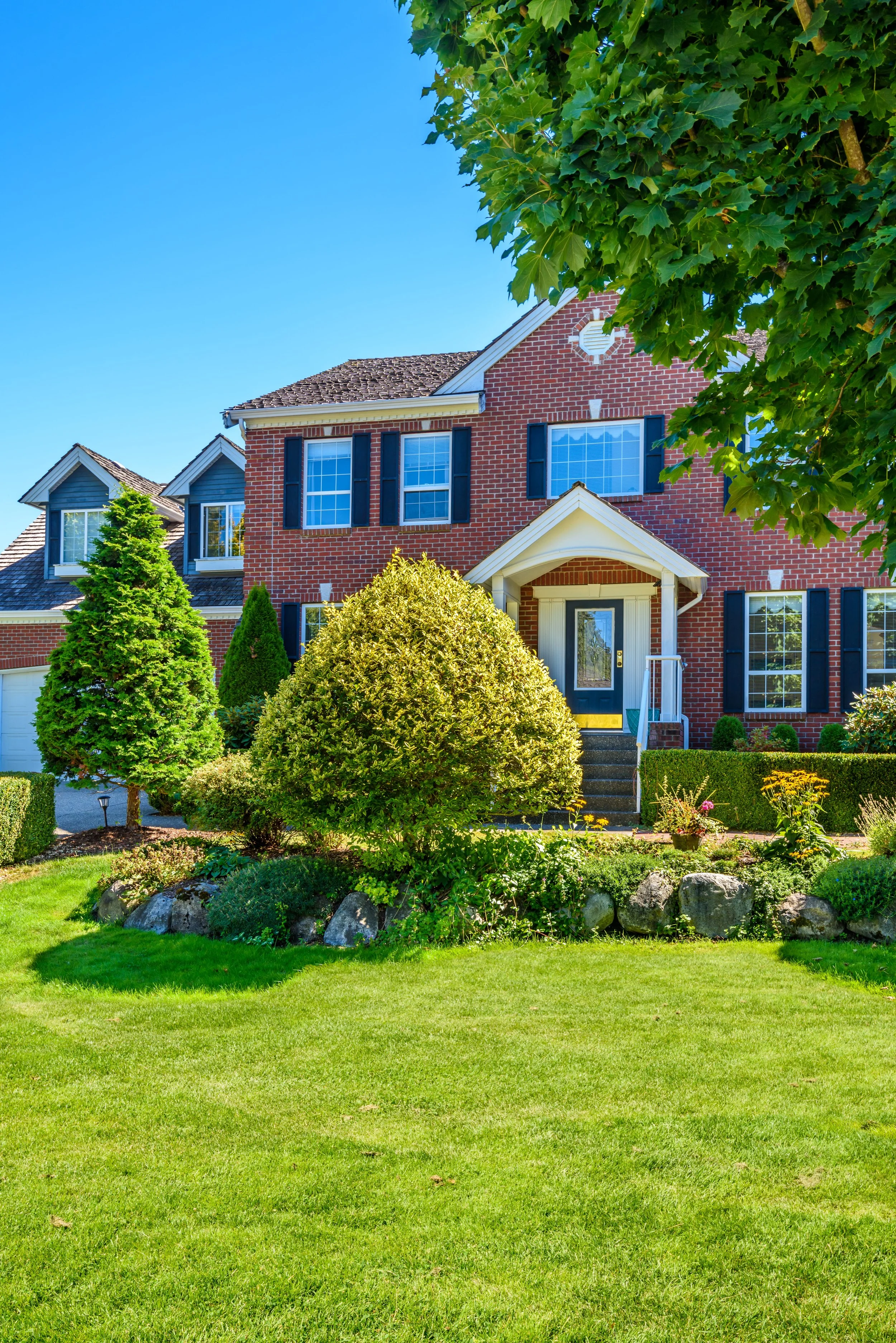 A brick house with black shutters, large windows, and a covered front porch, surrounded by well-maintained green lawn and shrubs, with trees in the yard and a clear blue sky.