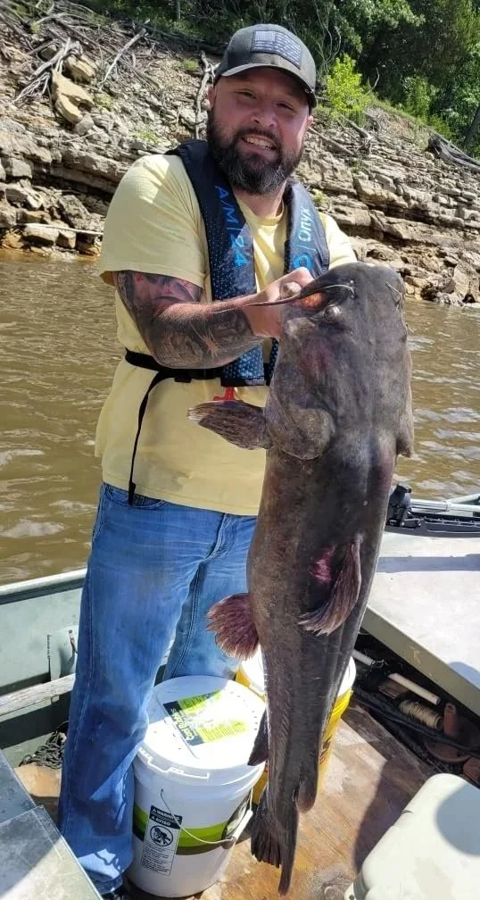 Man with a beard and tattoos on his arm, wearing a cap, yellow t-shirt, and a life jacket, holding a large catfish on a boat in a river with rocky shoreline and green trees in the background.
