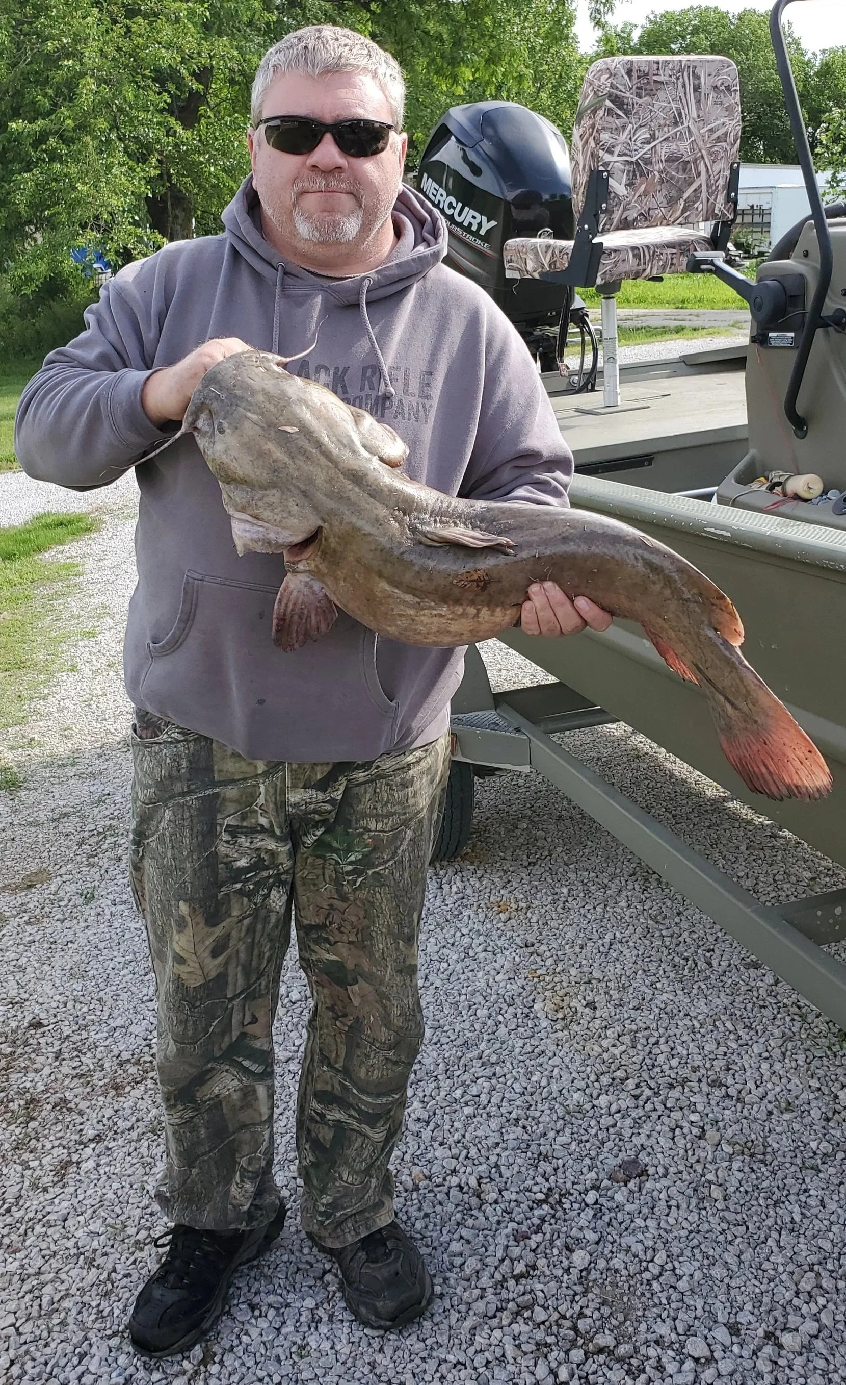 A man wearing sunglasses, a gray hoodie, and camouflage pants is standing outdoors on gravel, holding a large catfish and posing in front of a boat with a Mercury outboard motor and camouflage seat.