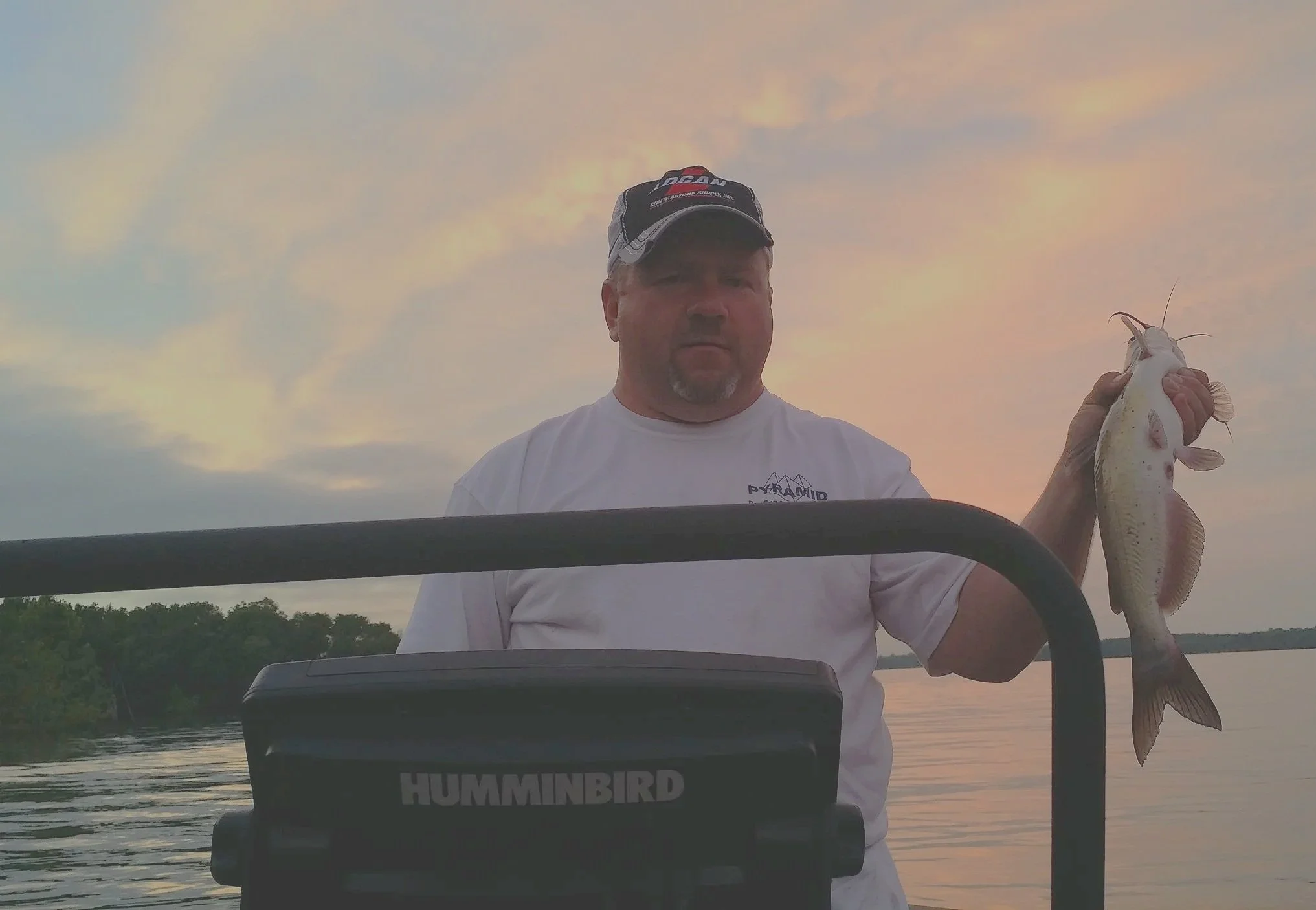 A man wearing a white t-shirt and a cap is holding a fish on a boat at sunset with a body of water and trees in the background.