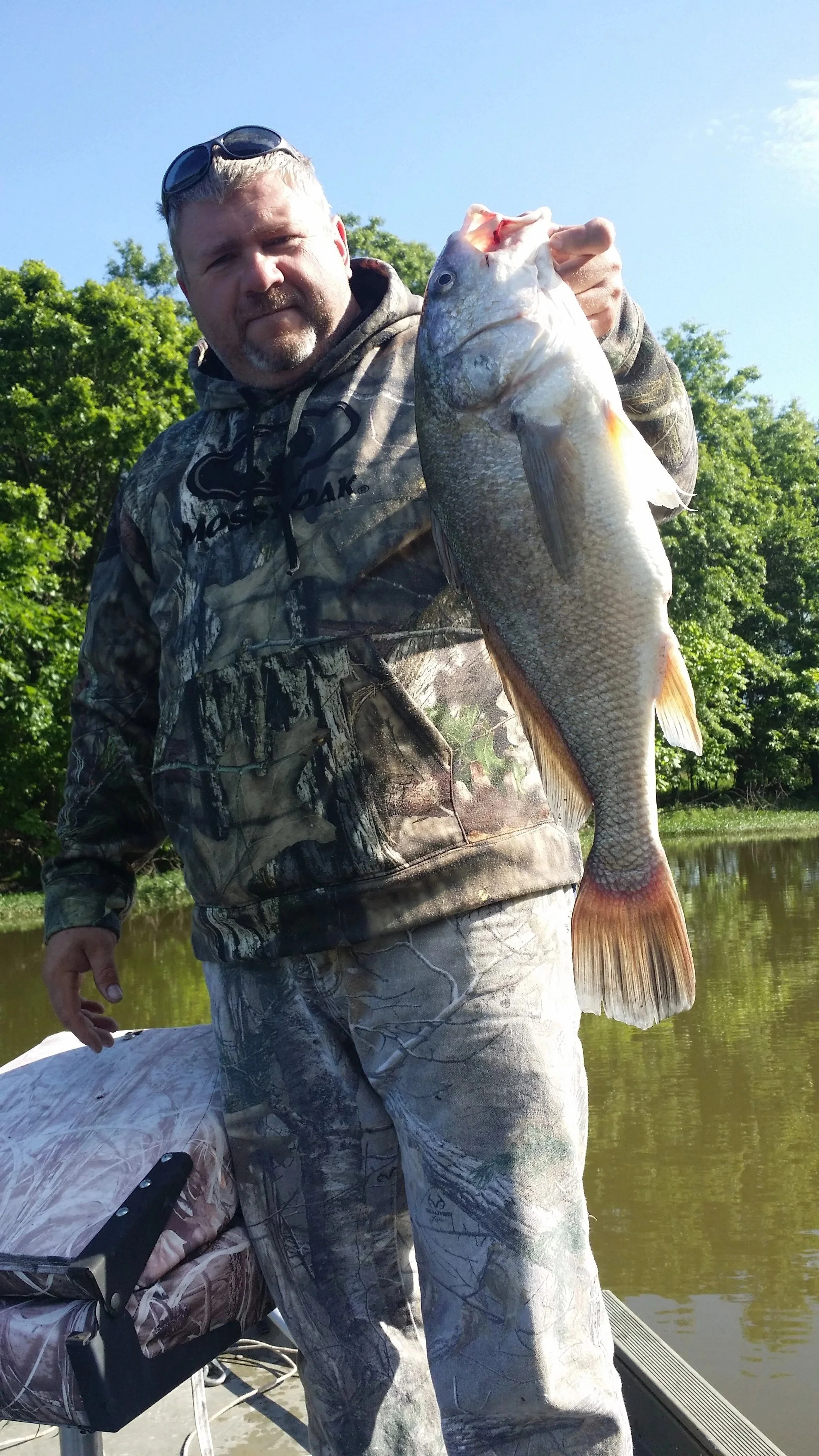 A man in camouflage clothing holding a large fish with water and trees in the background.
