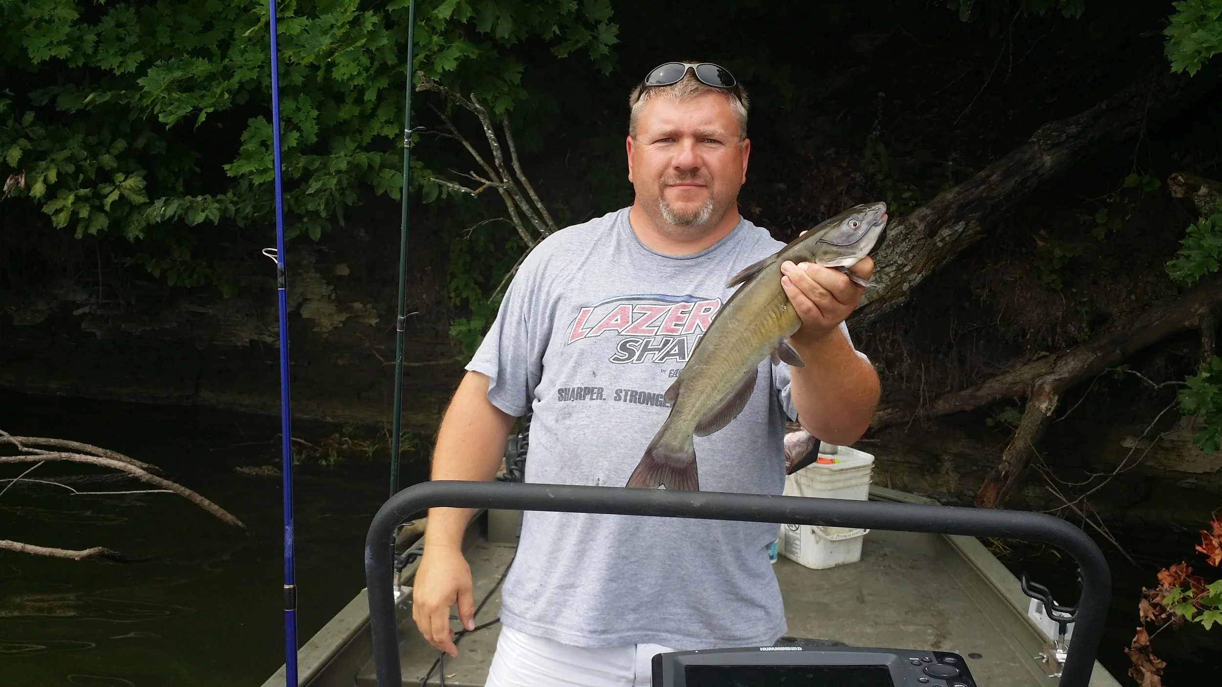 Man holding a fish on a boat with fishing rods and trees in the background