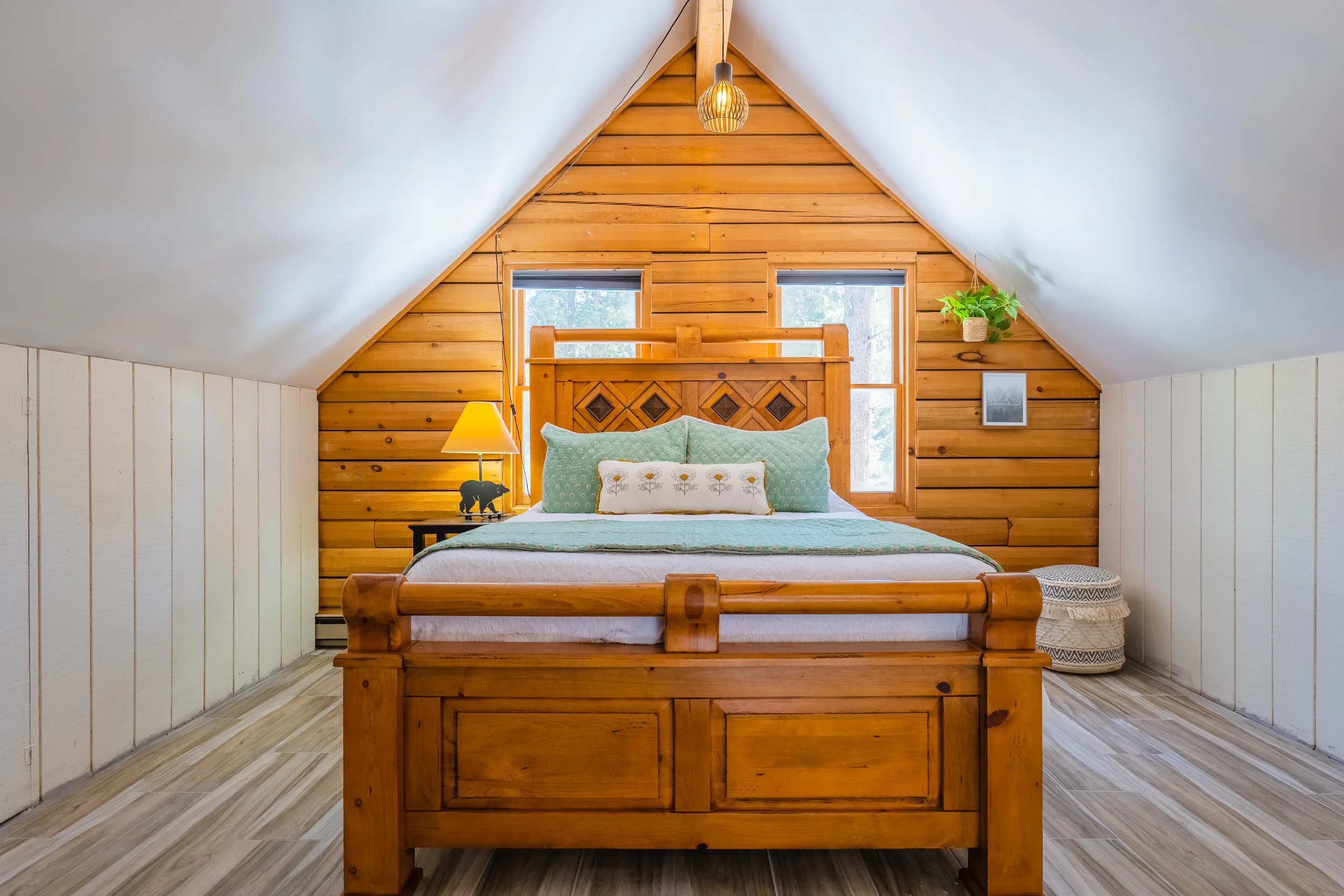 Living room with two dark leather sofas facing a fireplace with a stone surround and a mounted television. There are two side tables with lamps, a sliding glass door with curtains leading to a balcony, a wall clock, and a ceiling fan with lights. The room has wood flooring, a patterned rug, and a mountain landscape view outside.