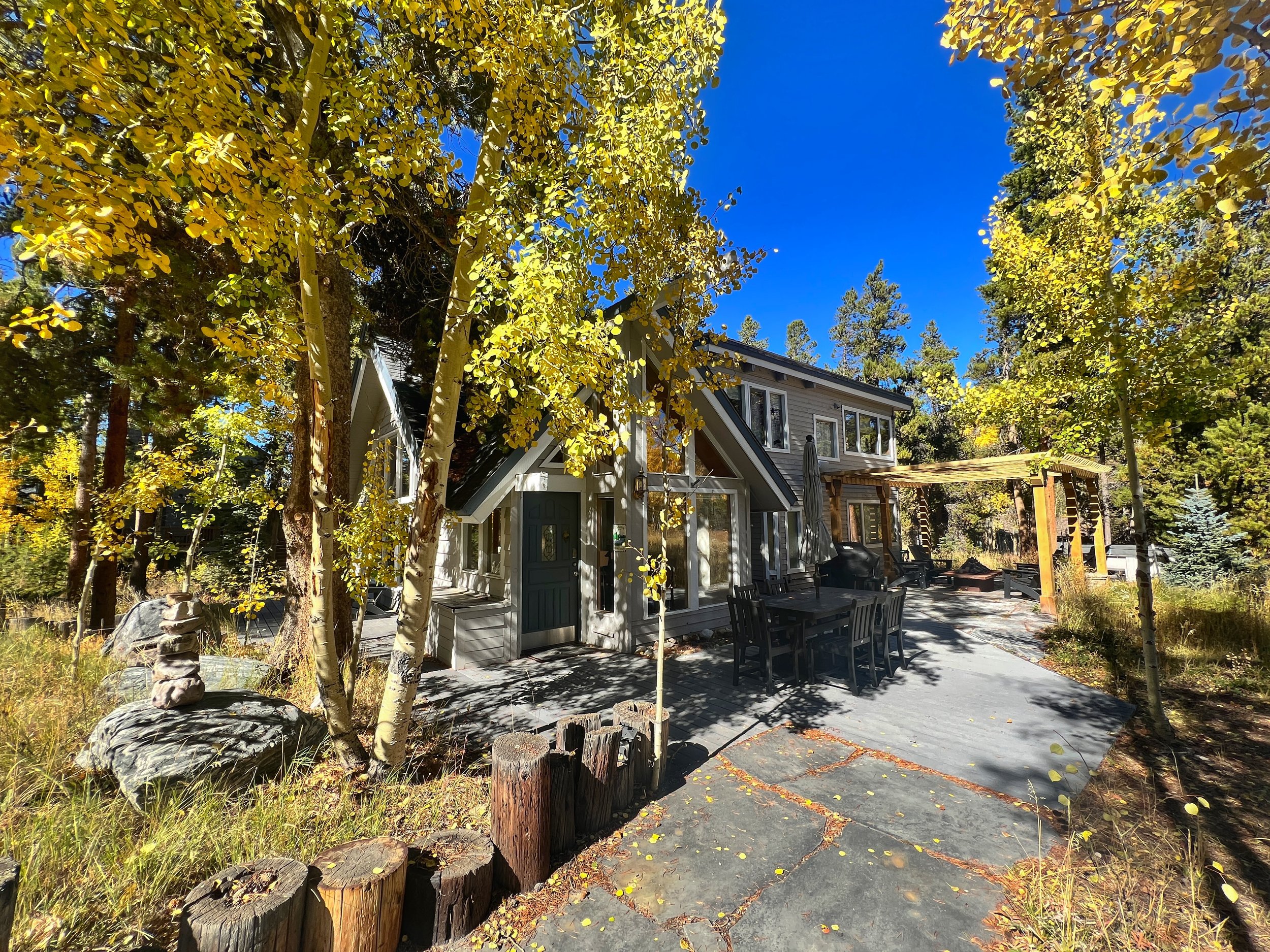 A two-story house surrounded by trees with yellow and green leaves, a paved patio with outdoor furniture, and an open wooden pergola, under a bright blue sky.