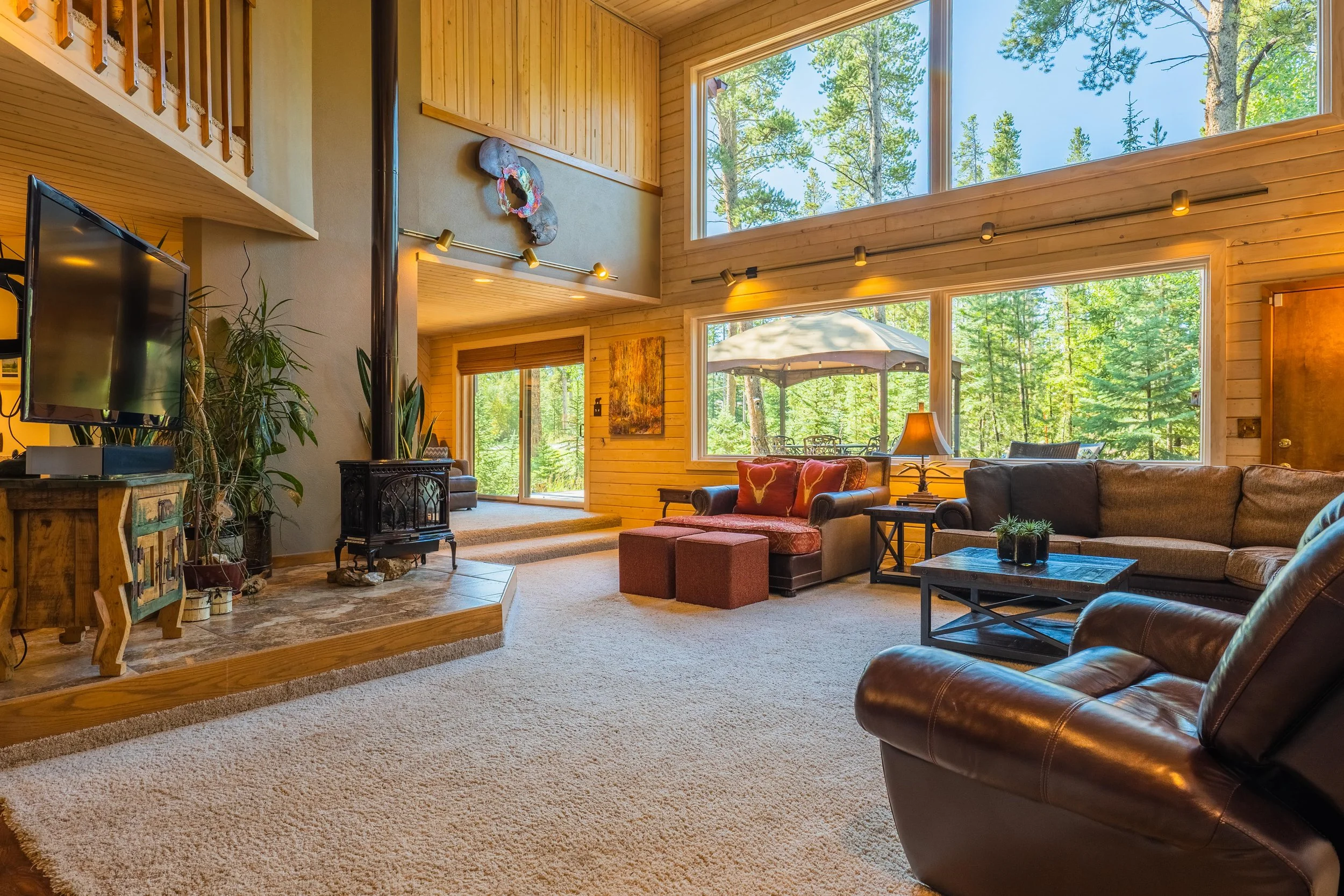 A spacious living room with large windows showing a wooded outdoor scene. The room features a brown leather armchair, a sofa, and an armchair with a floral pattern. There is a wood-burning stove, a television, and decorative plants. The walls and ceiling are made of light-colored wood, creating a cozy, rustic atmosphere.