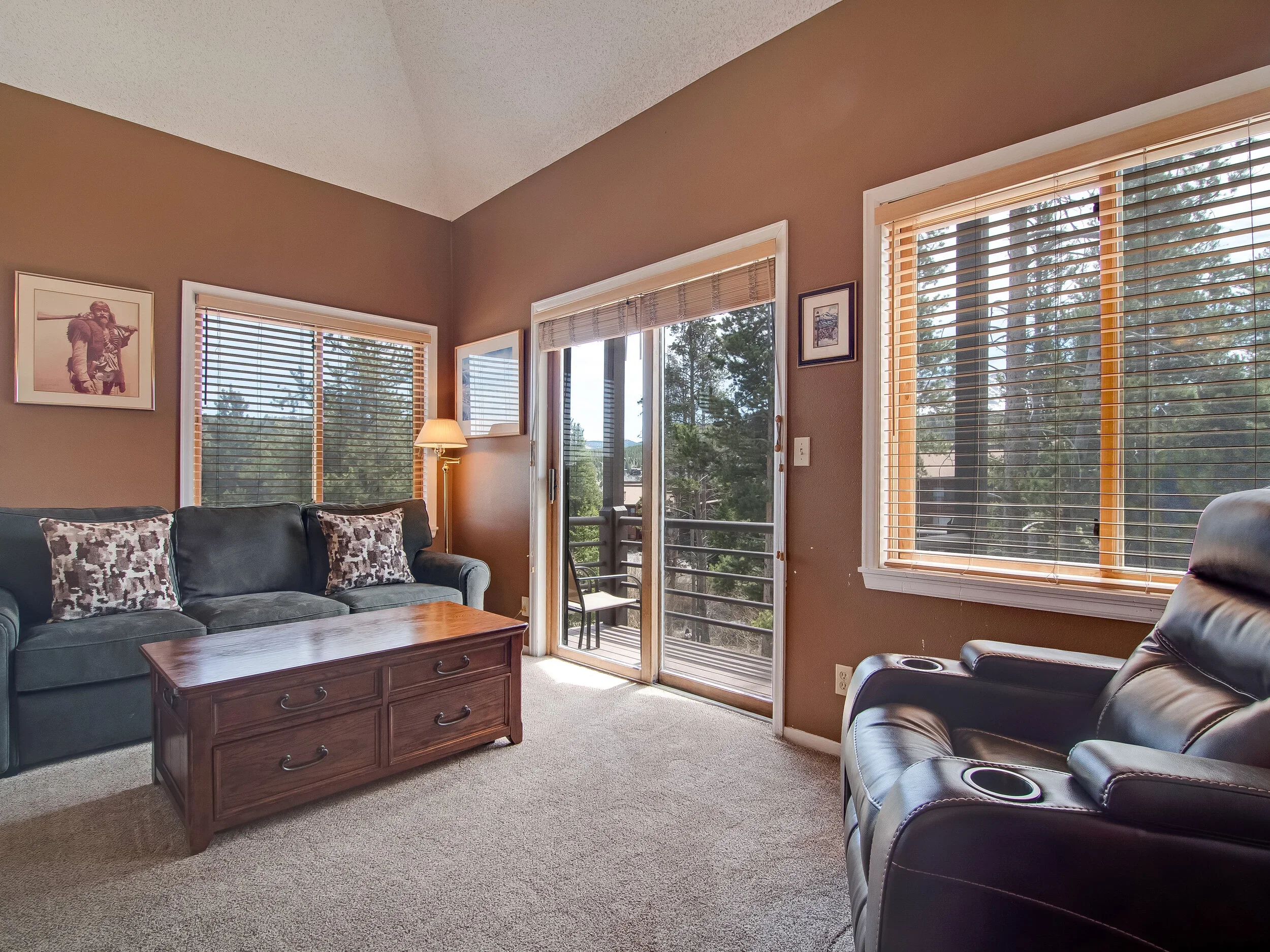 Living room with brown walls, three windows with blinds, a glass door to a balcony, a black leather recliner, a black sofa with patterned pillows, a wooden coffee table, a floor lamp, and framed pictures on the wall, overlooking trees outside.