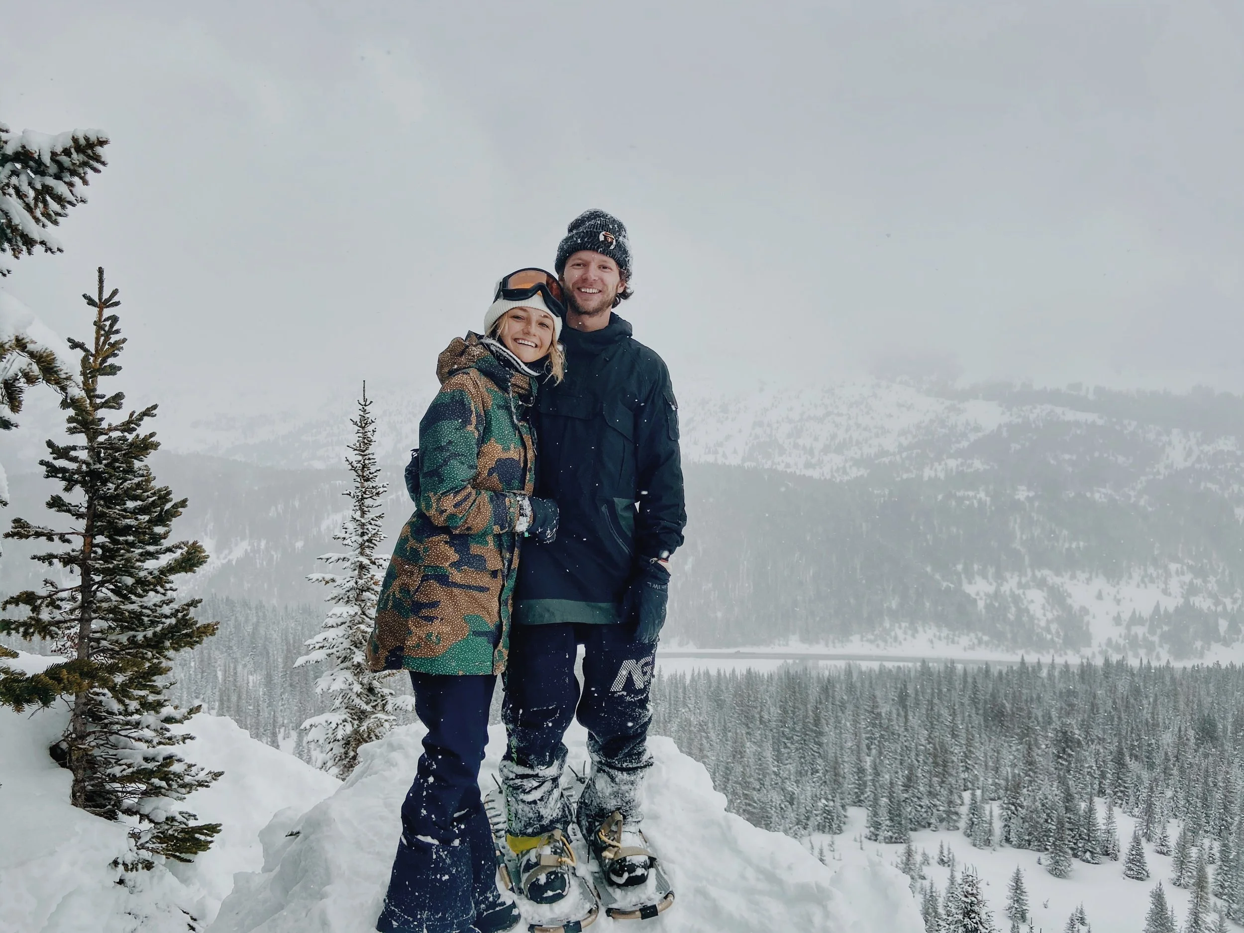 A smiling couple stands in the snow wearing winter clothing with snowshoe shoes, in a snowy mountain landscape with trees and fog in the background.