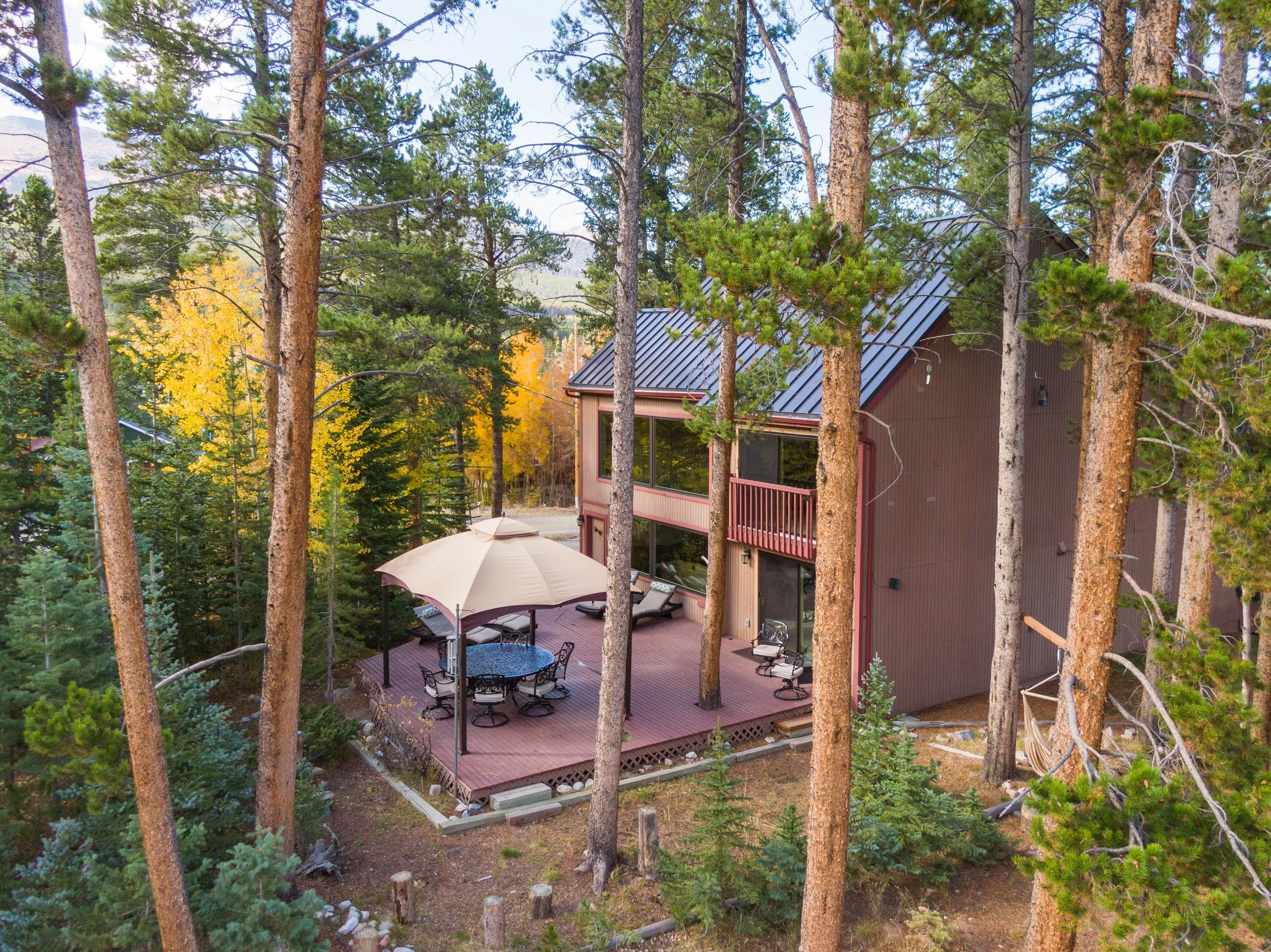A house in the woods with a large deck, outdoor furniture, umbrella, and surrounded by tall trees and yellow autumn leaves.