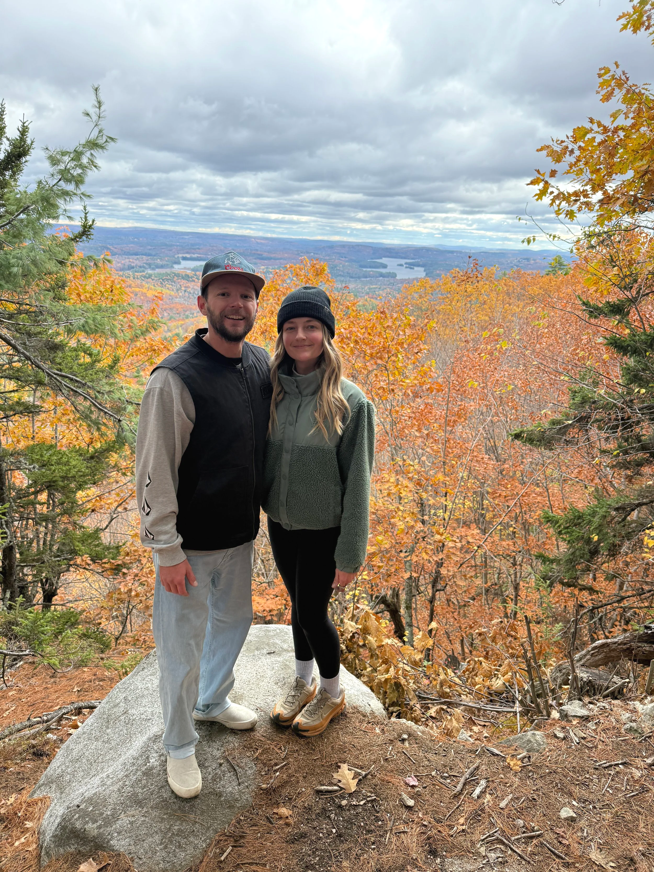 A man and a woman standing on a large rock in a forested area with vibrant autumn leaves, overlooking a scenic view of a lake and rolling hills under a cloudy sky.