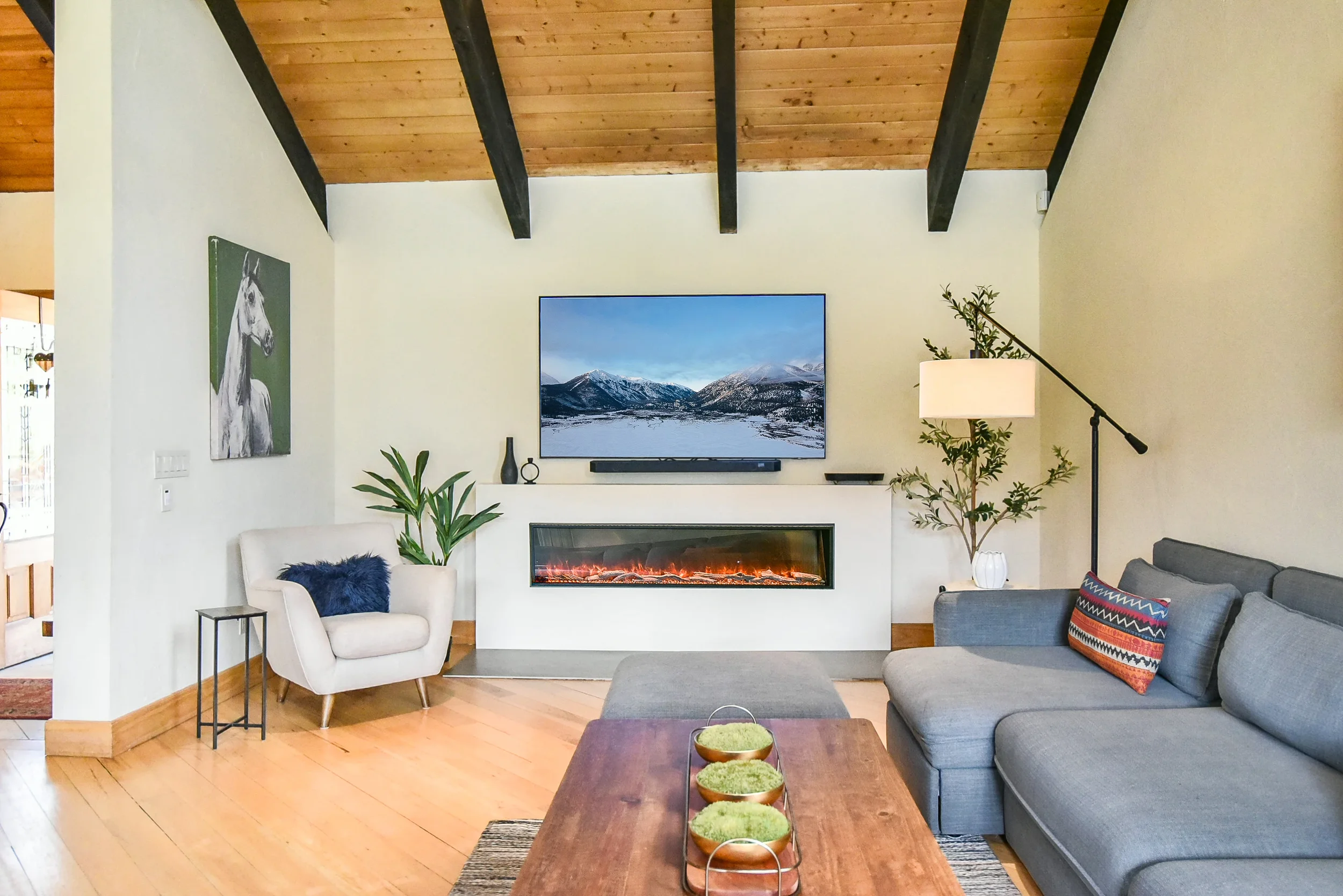 Living room with a black leather sofa, a gray armchair, a rattan chair with an orange cushion, various side tables, colorful pillows, large windows showing trees outside, and a door leading outside. The room has wooden accents and hanging light fixtures.