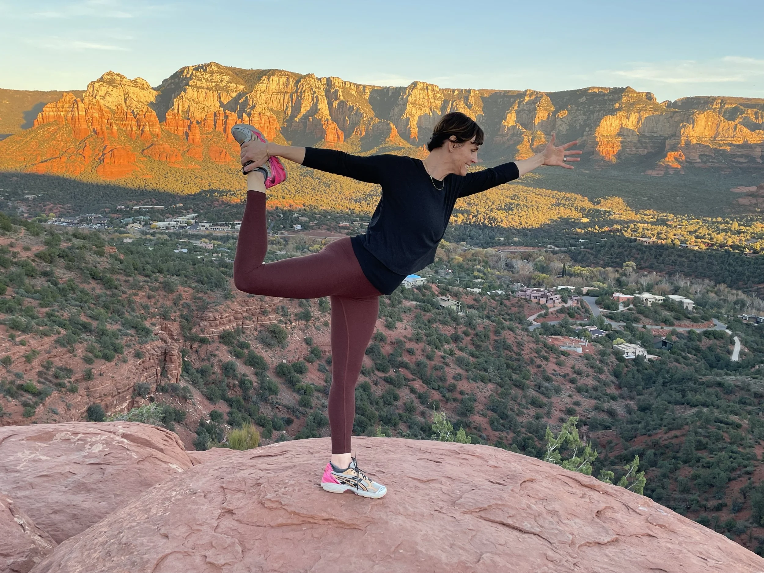 A woman practicing yoga on a rock ledge in a desert landscape with red rocks and mountains in the background during sunset or sunrise.