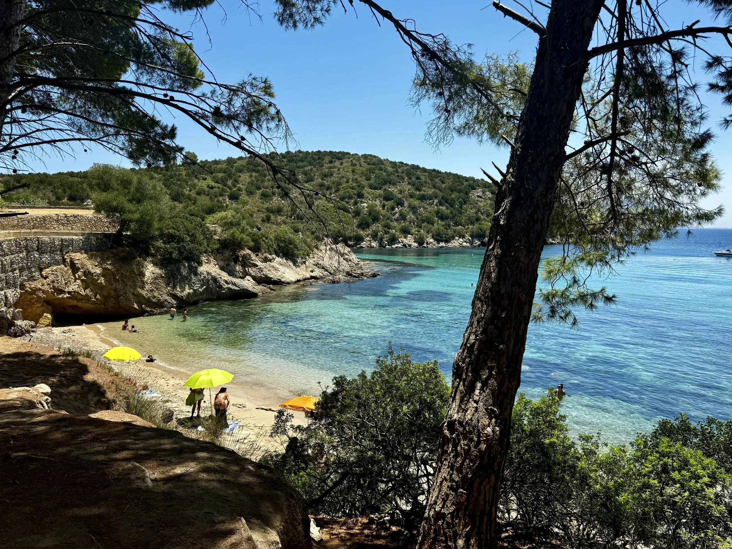 A scenic beach with clear turquoise water, surrounded by rocky cliffs and lush green trees, with people relaxing under yellow and orange umbrellas.