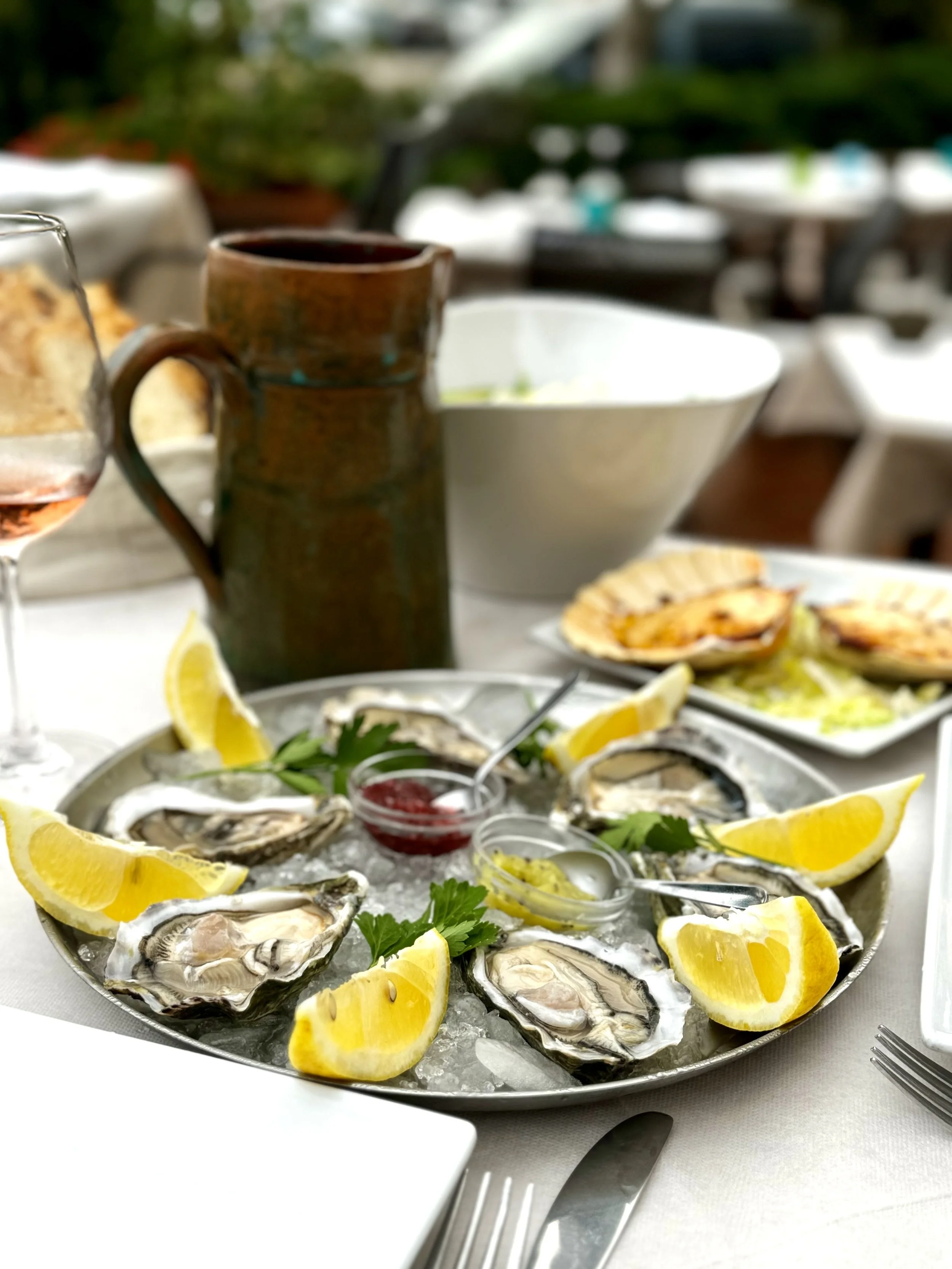 A seafood platter with fresh oysters, lemon wedges, cilantro leaves, and small containers of cocktail sauce and horseradish on a bed of ice, set on a white tablecloth at an outdoor dining area.