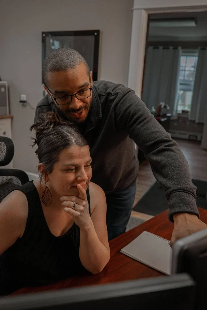 A man and woman look at a computer screen together, with the woman sitting at a table and the man leaning over her, in a home setting.