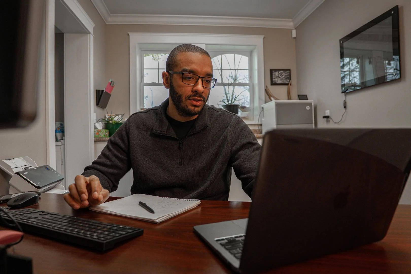 Man working at home on laptop, with notebook and pen, in a kitchen or dining area.