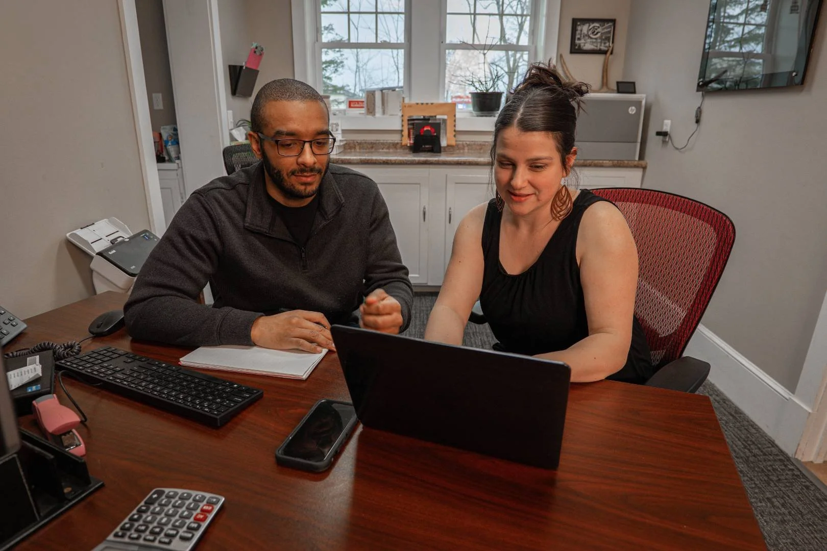 Two people sitting at a desk looking at a laptop, working together in an office or meeting room.