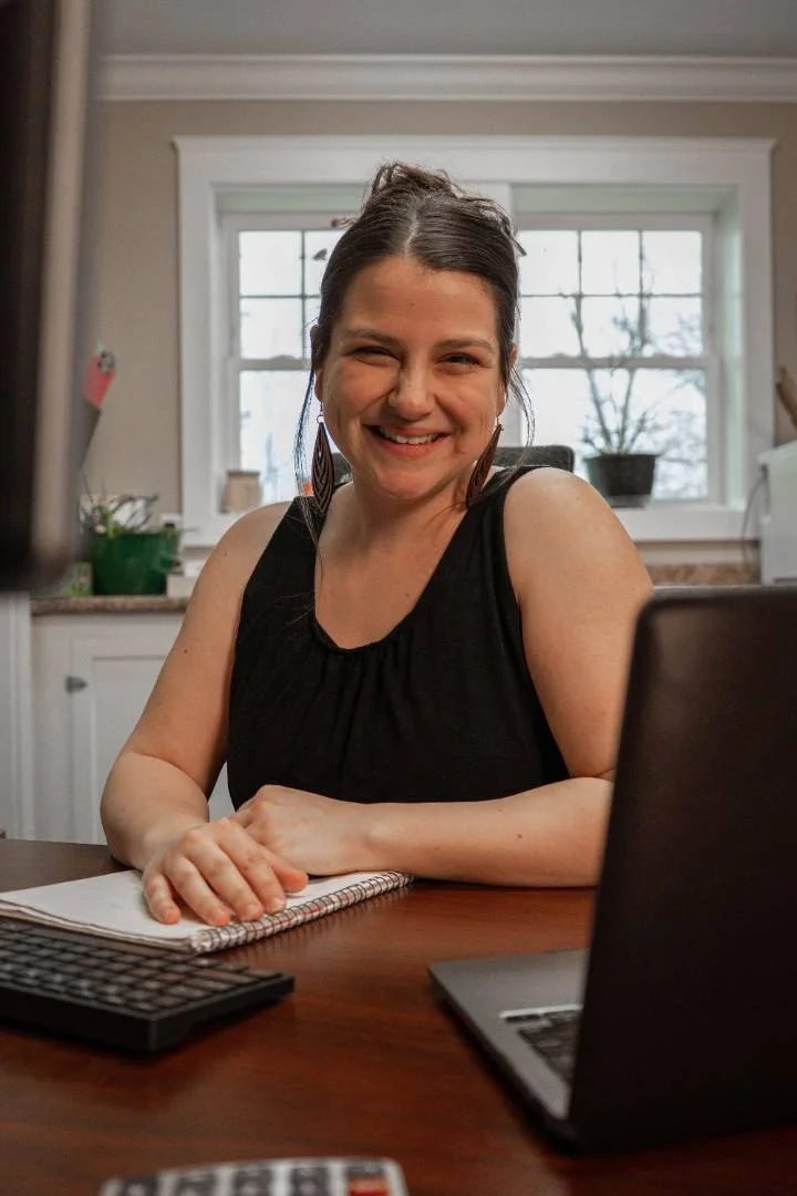 A smiling woman with dark hair in a ponytail, wearing a black sleeveless top, sitting at a wooden table with a notebook, remote, and open laptop, in a kitchen with a window and potted plant in the background.