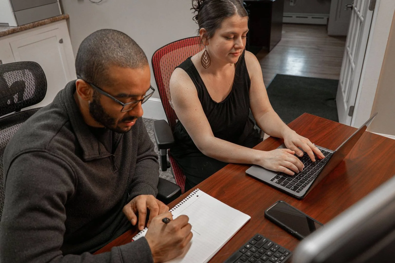 Two people sitting at a wooden table working on laptops and taking notes.