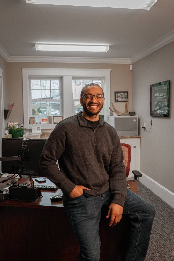 Smiling man in glasses and dark sweater standing in an office with a desk and windows behind him.
