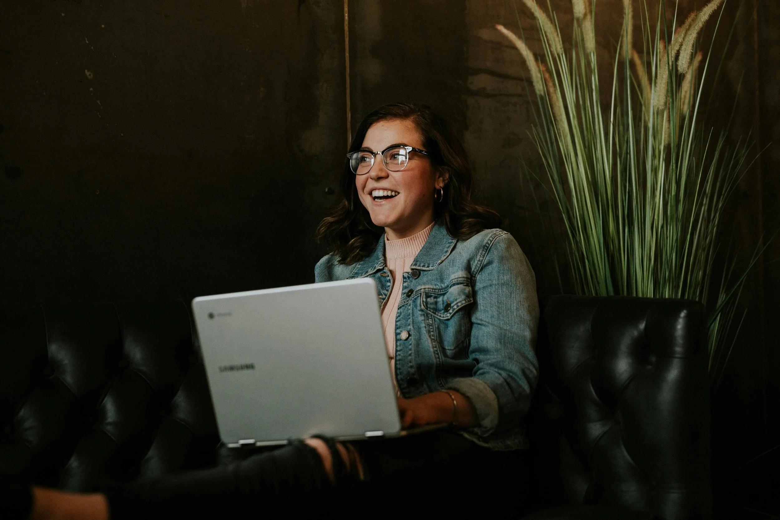 Woman using a laptop while seated on a leather sofa beside tall plants.